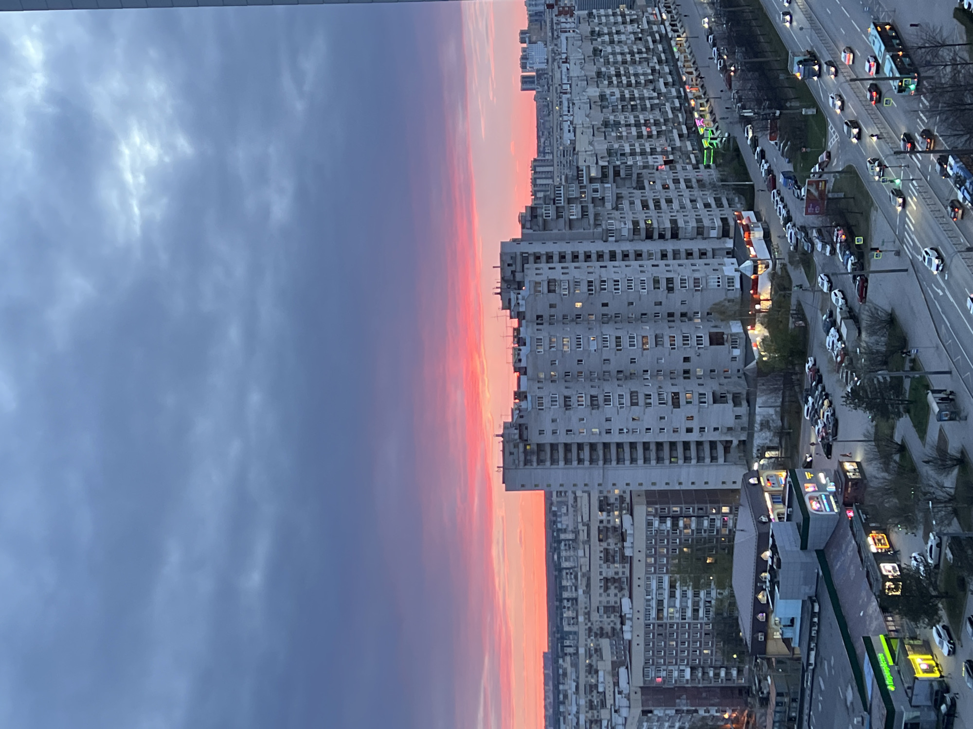 This is an elevated view of an urban landscape in Saint Petersburg, Russia, captured during dusk or twilight. The image depicts a wide panorama with a dramatic sky and a bustling city below.
The sky occupies the upper portion of the frame, showcasing a striking transition of colors. Near the horizon, the sky glows with intense pinks, oranges, and soft reds, characteristic of a sunset. This vivid band gradually fades into cooler shades of grey and deep blue higher up, with scattered, diffuse clouds adding texture.
Below the horizon, the city stretches densely. Numerous multi-story residential buildings, primarily in the style of concrete slab high-rises, dominate the middle ground and foreground. Many windows in these buildings are lit, suggesting inhabitants or activity inside.
To the right, a major multi-lane road curves through the scene, busy with traffic. Several cars are visible, both moving and parked, along with at least two buses, one of which appears blue and white. Streetlights and vehicle headlights/taillights illuminate the thoroughfare, creating streaks of light and bright spots. Pedestrian crossings and traffic lights are also visible.
Along the road and at ground level, various commercial establishments are present, marked by illuminated signs. One prominent yellow and green sign displays the text "Перекрёсток," indicating a Perekrestok supermarket, a common chain in Russia. Other smaller commercial signs and kiosks add to the urban vibrancy. Some patches of greenery, including trees and grass, can be seen along the roadside and between the buildings, providing a contrast to the concrete structures.
The overall impression is one of a vast, active city transitioning from day to night, with the natural beauty of the sunset sky juxtaposed against the illuminated human-made environment.
