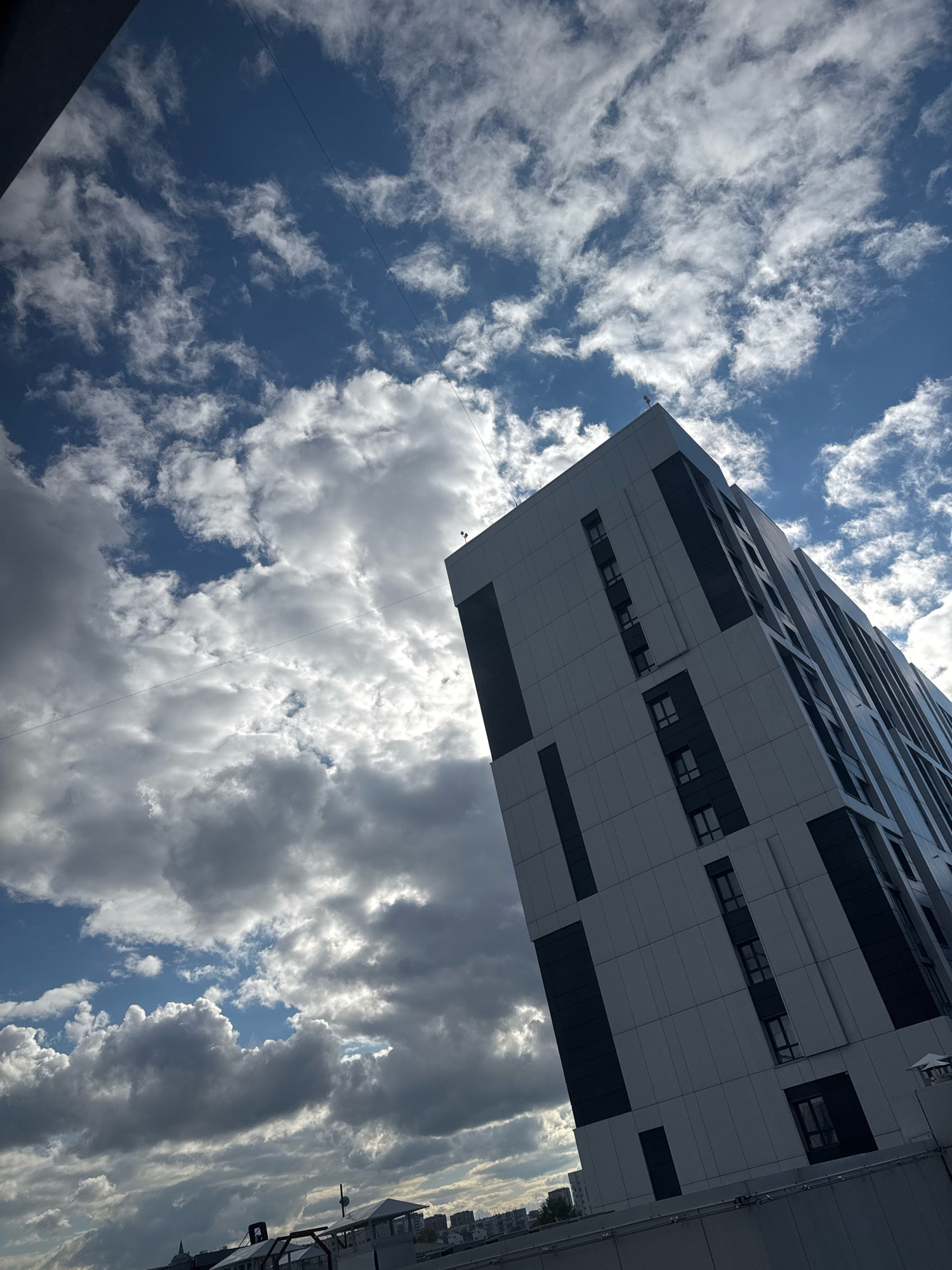 The image depicts a modern, tall residential building in Novosibirsk, Russia. The building has a light-colored facade with dark rectangular window panels. The perspective is from below, looking up at the building against a dramatic sky filled with white and grey clouds. The sky is a deep blue in some areas, with sunlight breaking through the clouds in others. The building is angled in the frame, giving it a dynamic appearance. In the foreground, a rooftop railing is partially visible. The overall scene suggests an urban environment with a focus on architectural design and the interplay of light and shadow on the building and sky.