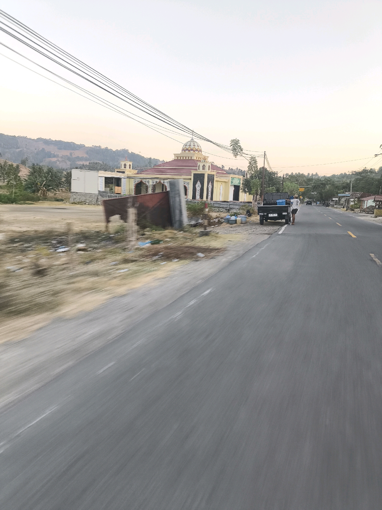 A low-angle, motion-blurred medium shot captures a roadside scene in Padengo, Indonesia, as viewed from a moving vehicle. In the mid-ground, a pale yellow mosque with a red-tiled roof and a prominent dome stands to the left of a paved road. Electrical wires crisscross the pale sky above. A dark-colored pickup truck is parked on the shoulder next to the mosque, with a person wearing a white shirt and dark shorts standing beside it. The foreground consists of a dusty, dry embankment alongside the road, showing evidence of scattered debris. The road itself is asphalt, with yellow lane markings. In the background, rolling hills covered in vegetation and scattered trees recede into the distance under a hazy sky.