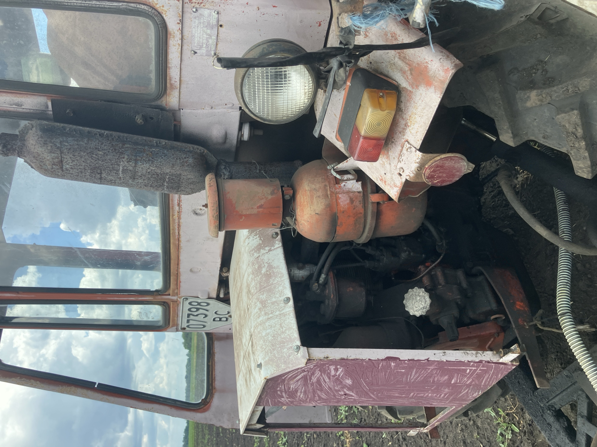 An aged, light-purple tractor is parked on a dark, muddy field under a cloudy sky. The tractor features a metallic exhaust pipe, a round headlight, and a set of red and amber turn signals. A license plate with the number "07398 BC" is visible on the side of the cabin. The tractor's engine compartment is partially open, revealing various mechanical components. The surrounding environment is agricultural, with fields stretching into the background. The image appears to have been taken in Berdyansk, Ukraine, given the context provided.