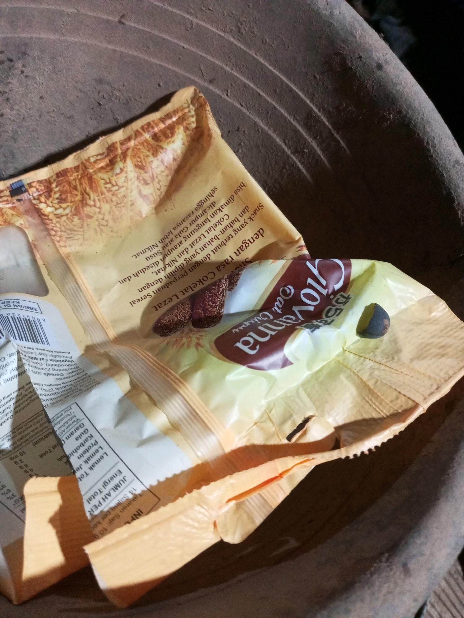 The image shows a close-up of an opened packet of "Yovanna Oat Crisps" lying in a brown, textured, bowl-like object. The packet is made of foil and paper, with a prominent "Yovanna" logo in maroon and white. Inside the packet, there are several crisp snacks visible, along with what appears to be a dried date or similar small, dark fruit. The background is dark and out of focus, with the primary focus on the food product. The location is described as Keling Panggau, Indonesia.