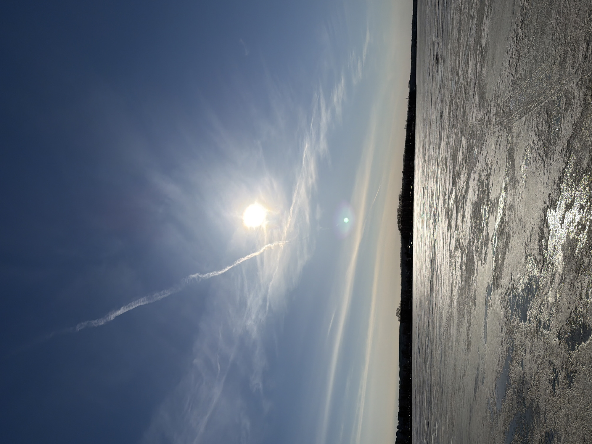 The image depicts a scene of a frozen body of water, presumably a lake or a river, under a bright, sunlit sky. The sky is blue with wispy clouds, and a contrail is visible near the sun. A shoreline with trees is visible separating the sky from the frozen water. The frozen surface has a textured appearance, reflecting the sunlight. The city of Votkinsk, Russia, is not explicitly identifiable within the scene. No people or specific objects are discernible in the image.