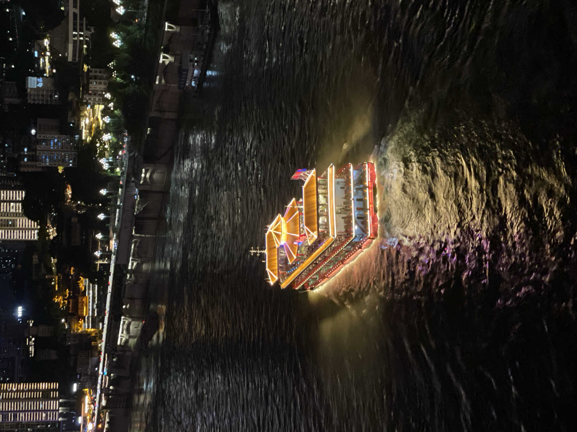 An elevated, high-angle night-time shot captures a brightly lit, multi-tiered tour boat navigating a wide, dark river. The boat, featuring traditional East Asian architectural elements with orange-tiled roofs and red and white siding, is illuminated by numerous lights, casting a warm glow on the water. The boat is creating a wake behind it. In the background, the Yuzhong District of Chongqing, China, is visible. The cityscape is characterized by tall, modern buildings with illuminated windows, some displaying Chinese characters. A long, elevated structure, possibly a bridge or embankment, with streetlights and some traffic, runs along the riverbank. The dark water of the river reflects the lights from both the boat and the city, creating a vibrant contrast.
