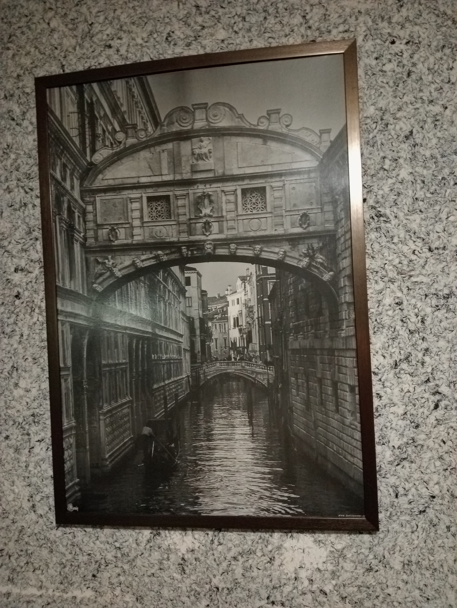 A black and white photograph depicts the Bridge of Sighs in Venice, Italy. The bridge is a striking architectural element, spanning a canal and connecting the Doge's Palace to the new prison. The photo captures the intricate stonework of the bridge, with its enclosed passageway featuring small windows. Below the bridge, a dark canal reflects the muted light, with a gondola visible in the water. The surrounding buildings are characteristic of Venetian architecture, with tall windows and detailed facades. The overall atmosphere is one of historical grandeur and quiet reflection.