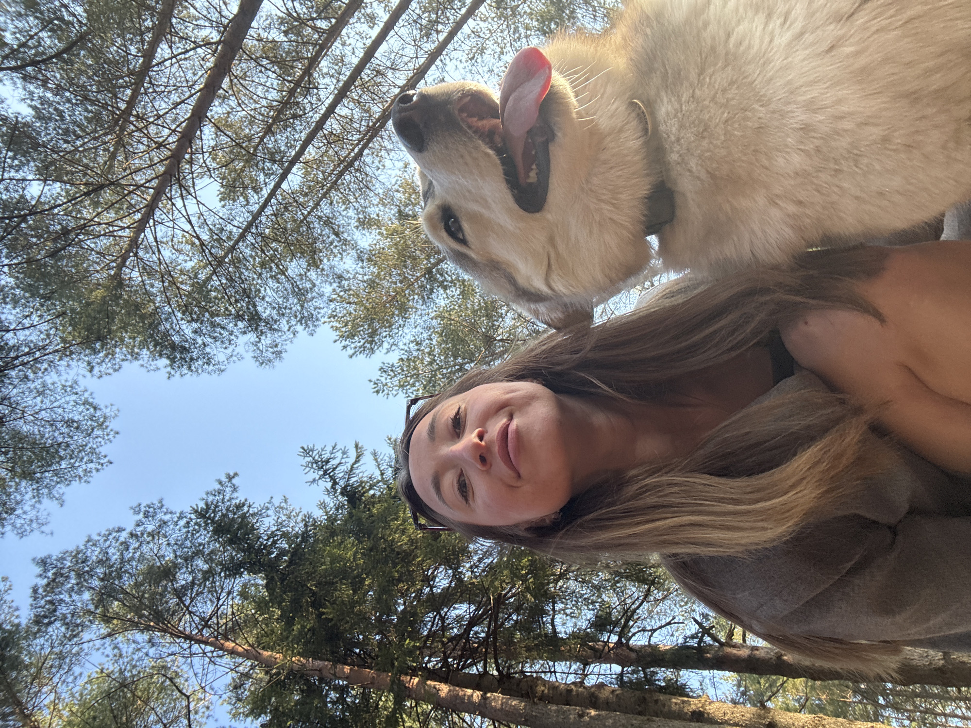 A woman and a dog are in a wooded area. The woman has long brown hair and is wearing sunglasses on her head. The dog is a large, light-colored breed with its mouth open and tongue out. The background features tall trees with green foliage and a blue sky. The image is taken from a low angle, looking up at the subjects. The exact location within Прикамский, Russia, cannot be determined from the image.