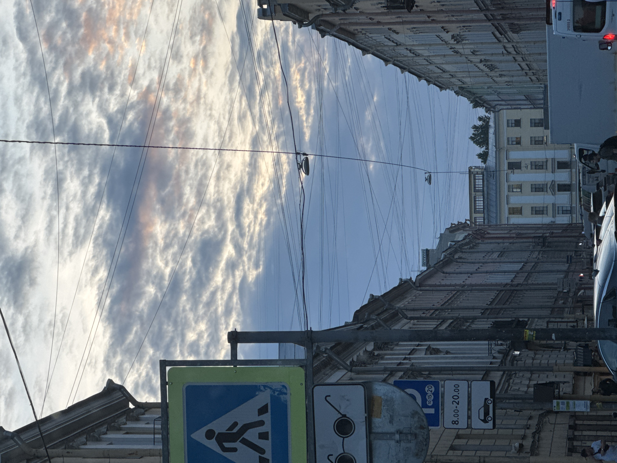 The image, displayed vertically, captures a view looking upwards and along an urban street in Saint Petersburg, Russia.
The sky, occupying the upper portion of the frame, is partly cloudy with patches of blue. White, textured clouds show soft pink and orange hues, suggesting either sunrise or sunset. A dense network of overhead utility wires and cables crisscrosses the entire sky, extending between the buildings on either side. A street light fixture is suspended from these wires in the upper-middle section.
Buildings with ornate, historical facades line both the left and right sides of the image, receding into the background. The building on the right has a distinct cream-colored facade with many windows.
At the bottom of the image, street-level elements are visible. On the left, a bright blue pedestrian crossing sign with a white walking figure is prominent, bordered in yellow. Next to it, a metallic round sign is partially obscured, and then a white rectangular sign clearly displaying the text "8.00-20.00". Adjacent to this, another white rectangular sign with a black car icon, and a blue circular sign with "10 15 20". On the far right, parts of a white delivery van are visible, with "21535 2" faintly discernible on its rear. Another darker vehicle is partially seen further into the scene. An indistinct human figure is vaguely visible on the right, near the white van.
The scene is a static depiction of daily life, showcasing the unique urban environment of Saint Petersburg, characterized by its classic architecture and the intricate overhead wiring infrastructure. The soft lighting on the clouds indicates either early morning or late evening.