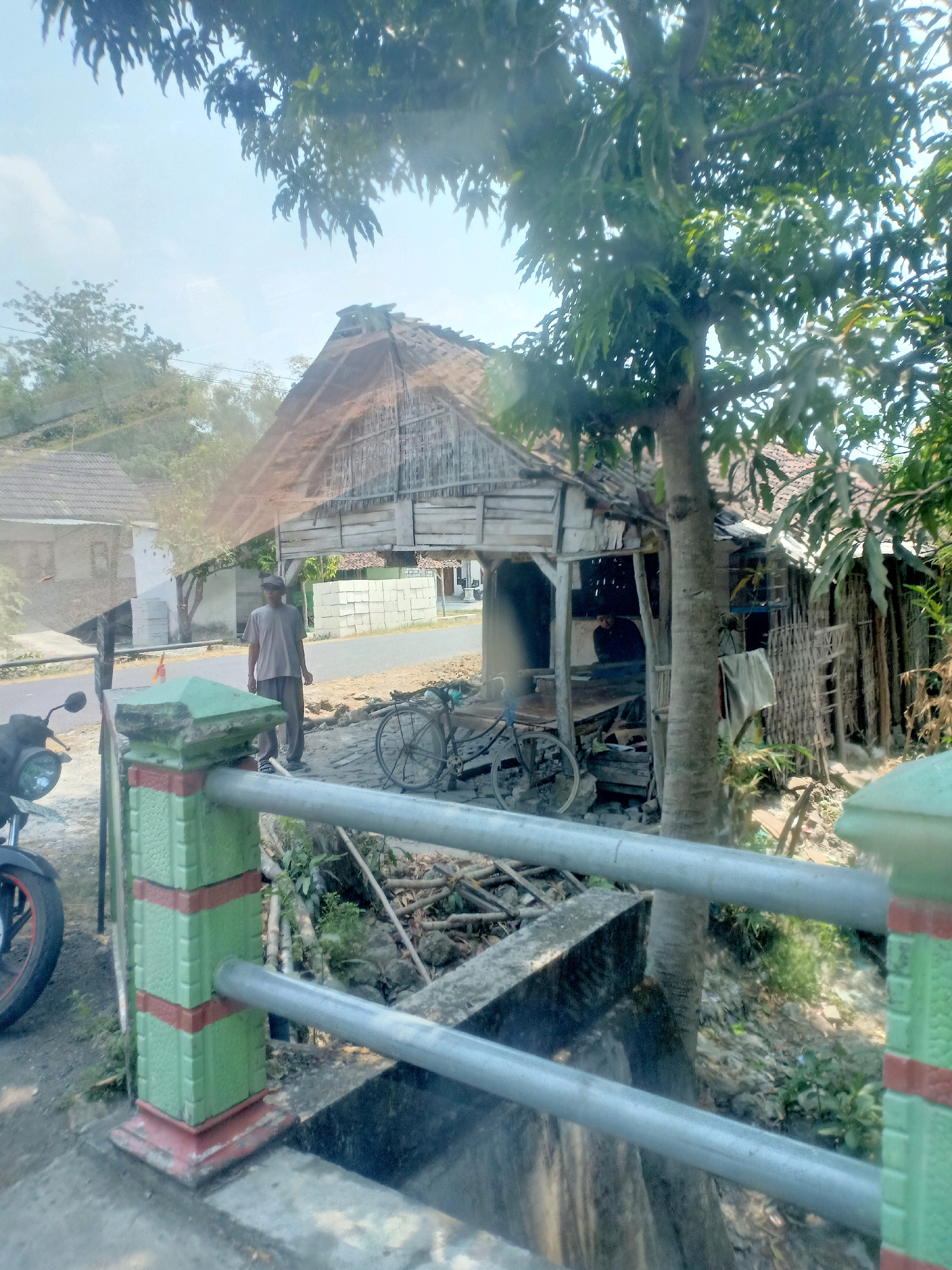 The image shows a rural scene in Turigede, Indonesia. In the foreground, a decorative fence post painted green with red accents stands next to a metal railing. Beyond the railing, a dirt path leads to a rustic dwelling with a thatched roof and bamboo walls. A man in a grey t-shirt and pants stands near the dwelling, and a bicycle is parked beside it. A motorcycle is visible on the left side of the image. The background consists of more dwellings, trees, and a clear sky. The overall impression is one of a simple, rural environment.