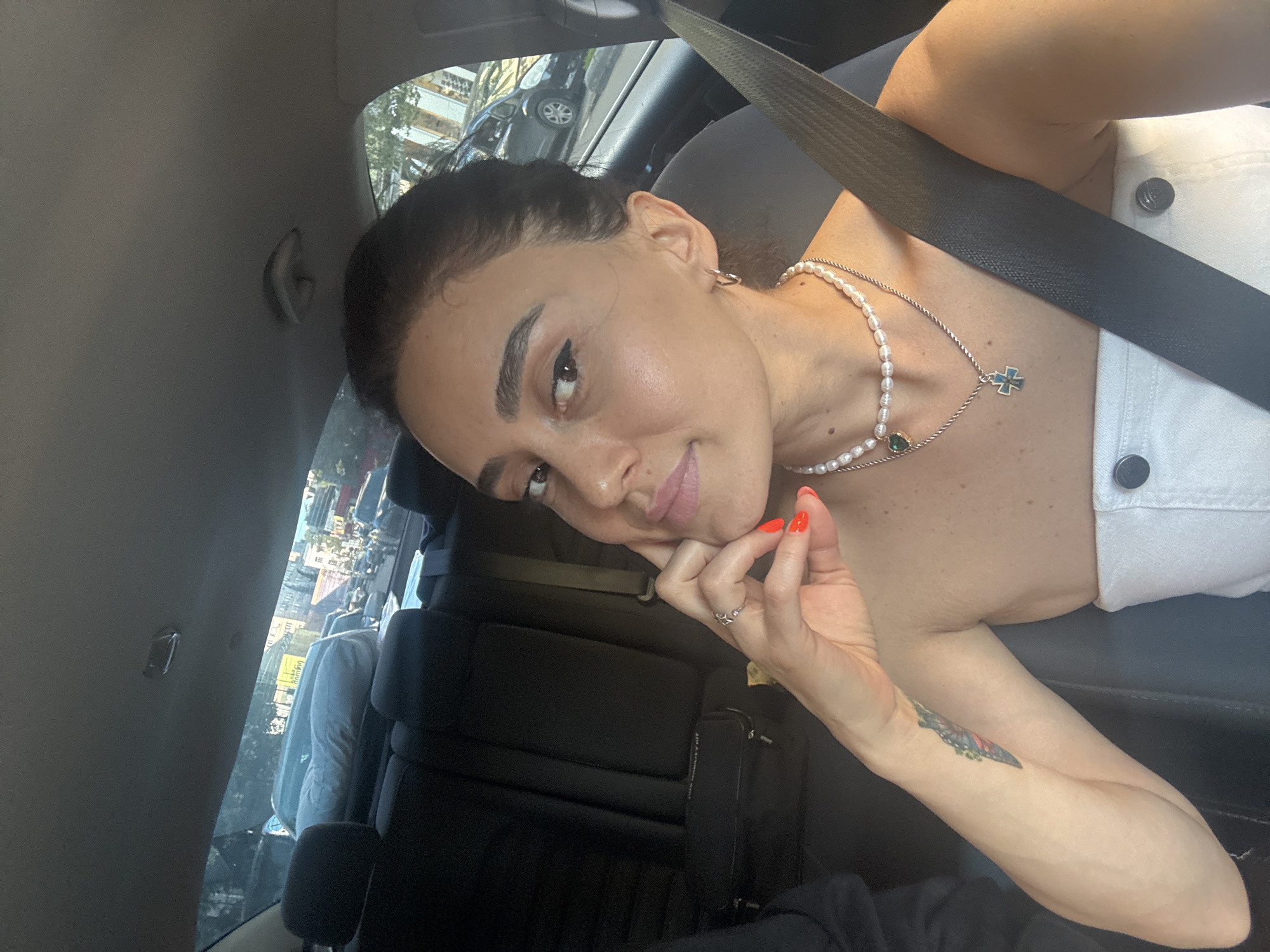 A medium shot photo, taken from inside a car, shows a woman looking towards the camera. She has dark hair pulled back, a light complexion, and dark eyes. Her left hand is raised to her chin, with bright orange nail polish visible. She is wearing a white strapless top and two necklaces: one pearl, and another silver chain with a cross pendant. A seatbelt is visible across her chest. In the background, through the car windows, there is a blurred view of a city street with buildings and other cars, consistent with a scene in Tbilisi, Georgia. The interior of the car shows black seats and a portion of the ceiling.