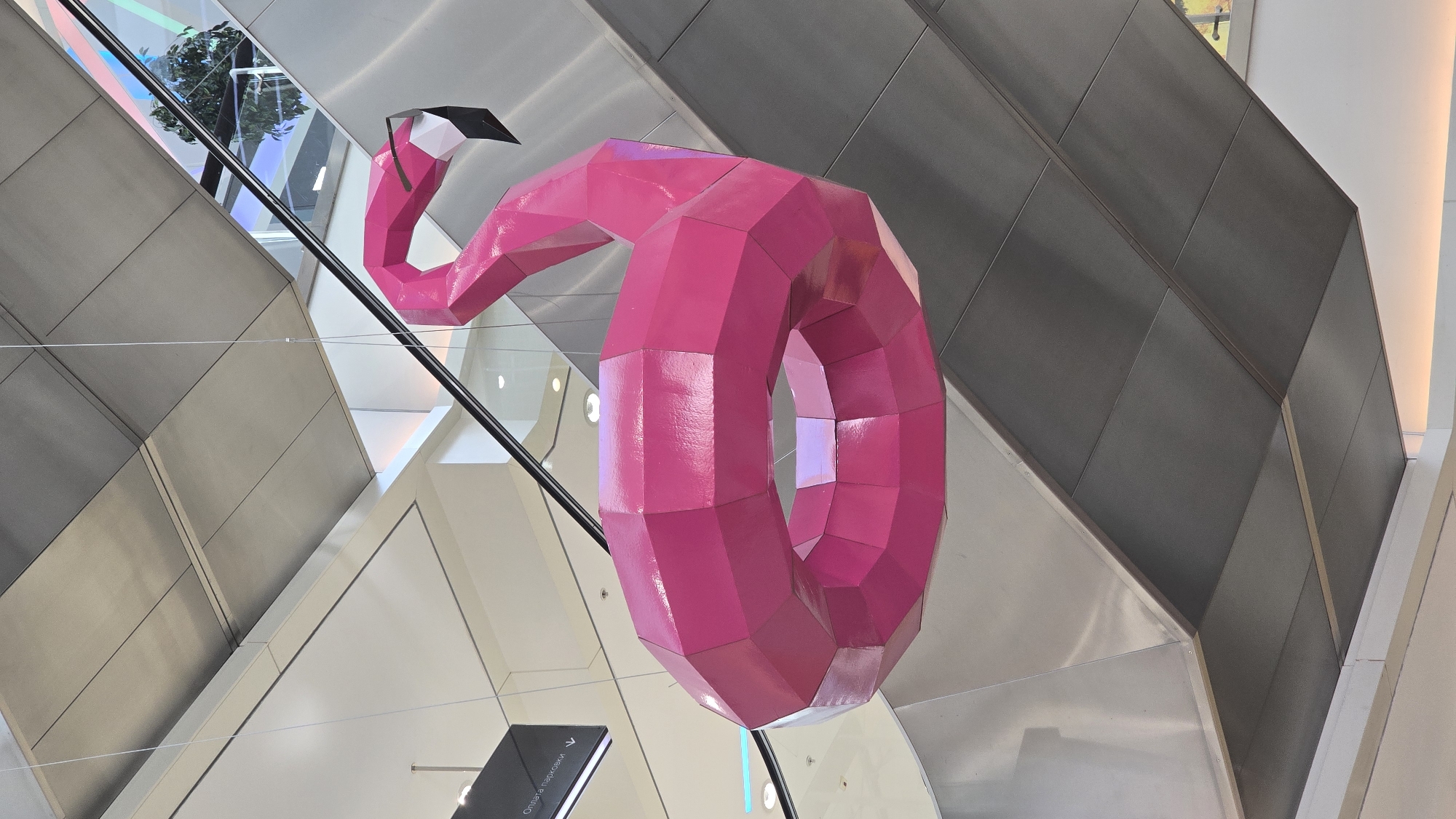 A large, polygonal, bright pink flamingo sculpture is suspended by thin wires from the ceiling of an indoor space, possibly a shopping mall or airport. The sculpture is abstract in design, with geometric shapes forming its body and neck, and it is curled in on itself to form a circular shape. The flamingo's head is a distinct feature, with a black tip on its beak. The background shows modern architecture with metallic and white surfaces, including escalators and what appears to be a ceiling with various panels and lighting. The overall impression is a contemporary interior space decorated with a vibrant art installation. The location is the Mall Metropolis in Khimki, Russia.
