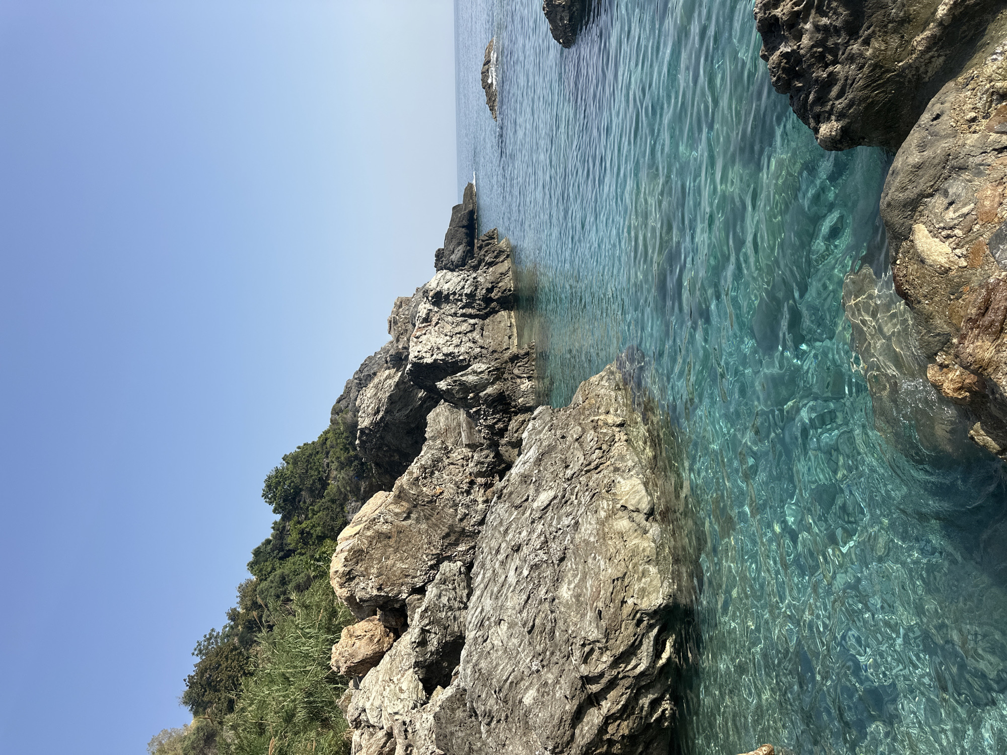 The image captures a coastal scene in Gazipaşa, Turkey, featuring a rocky shoreline meeting clear, turquoise water. The sky is a bright, cloudless blue. The foreground consists of large, grey and brown rocks of varying sizes, forming a rugged edge along the water. Vegetation, including trees and grasses, grows on and around the rocks, suggesting a natural, undeveloped landscape. The water is remarkably clear, allowing visibility of the seabed beneath. The image is shot at an angle, likely from the shore looking out at sea.