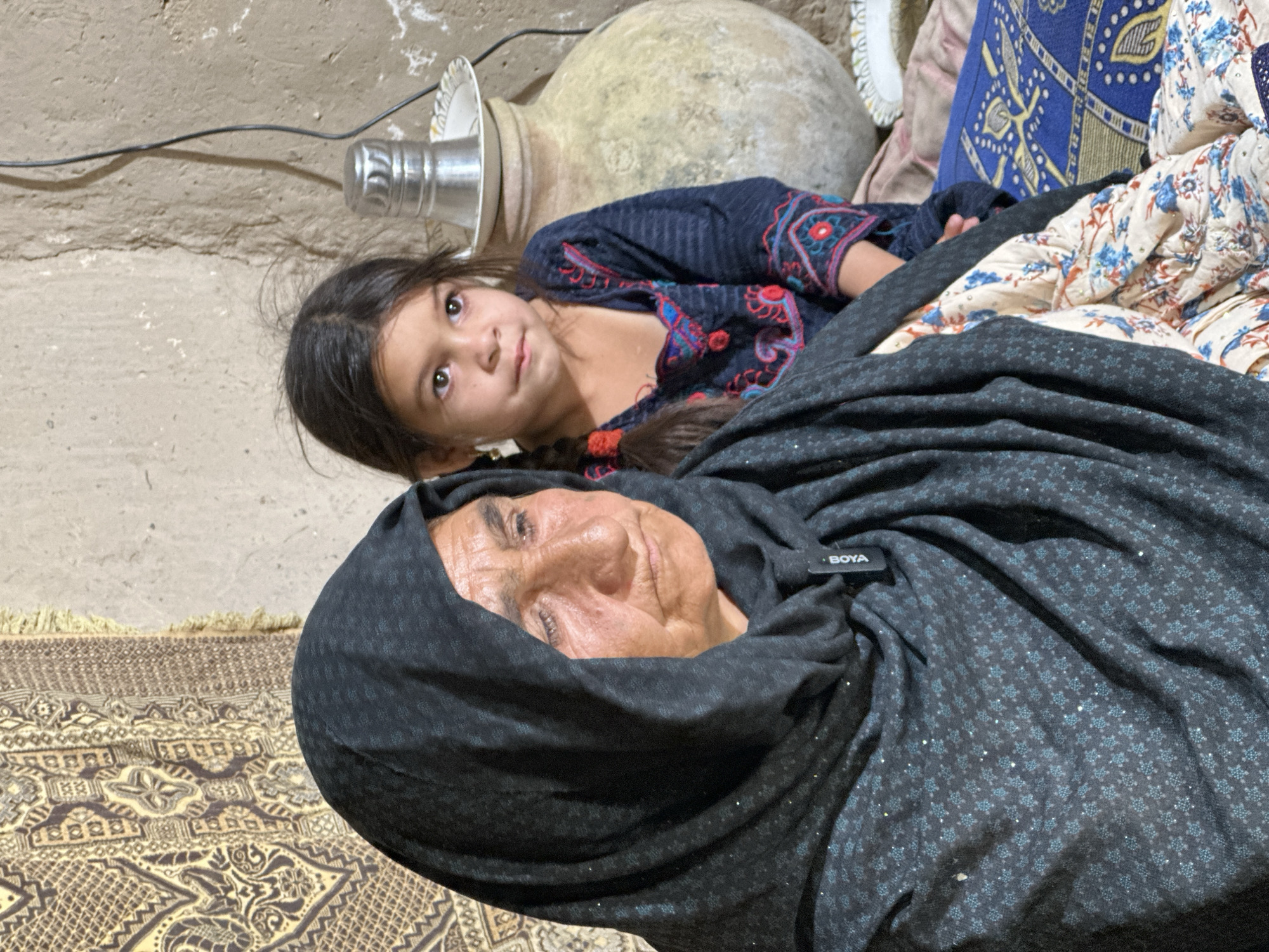 A low-angle shot captures an elderly woman with tanned skin, partially obscured by a dark, patterned headscarf or shawl, resting on a rug. Adjacent to her, a young girl with dark hair and eyes, wearing a dark blue top with colored embroidery, reclines. The setting appears to be indoors, with a beige concrete wall visible in the background. A ceramic pot and a metallic container sit near the girl. A patterned rug lies beneath them, and a colorful pillow or blanket is also visible. The location is Quetta City Tehsil, Pakistan.