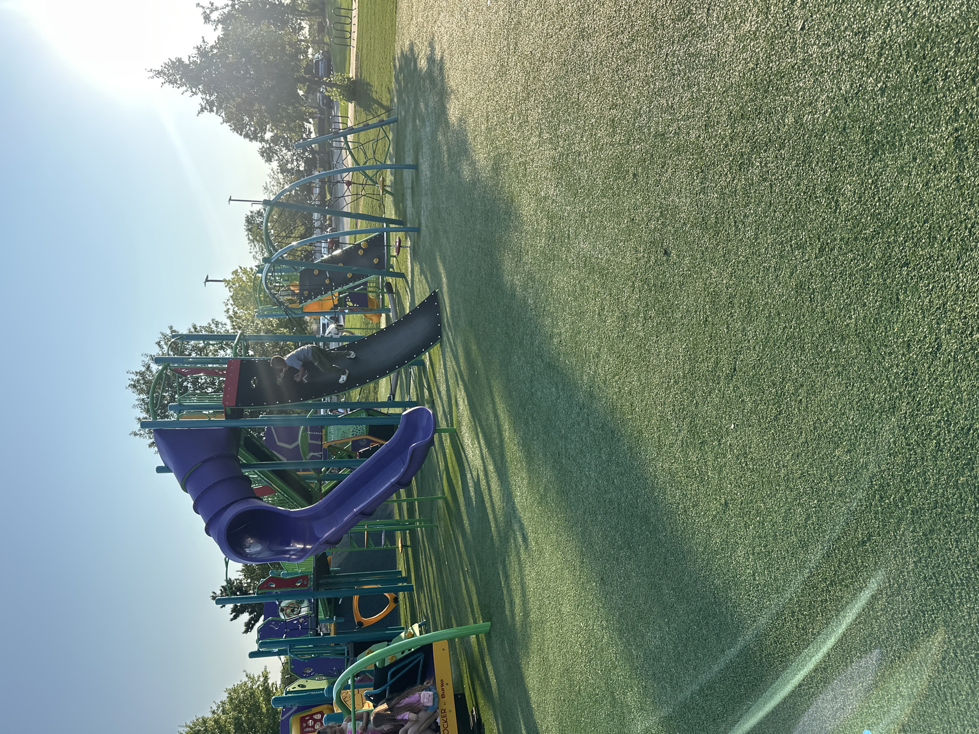 The image depicts a public playground on a sunny day. The playground features a large, multi-colored play structure with purple and black slides, climbing walls, and swings. Several children are visible on the playground, with one child actively climbing a slide and another appearing to be sitting on a swing. The ground is covered in artificial turf, which is bright green and shows the dappled shade of nearby trees. In the background, there are more trees, a parking lot with vehicles, and bicycle racks. The sky is a clear blue with the sun shining brightly, creating lens flare in the upper right portion of the image. The scene is located in Hoffman Estates, United States of America.
