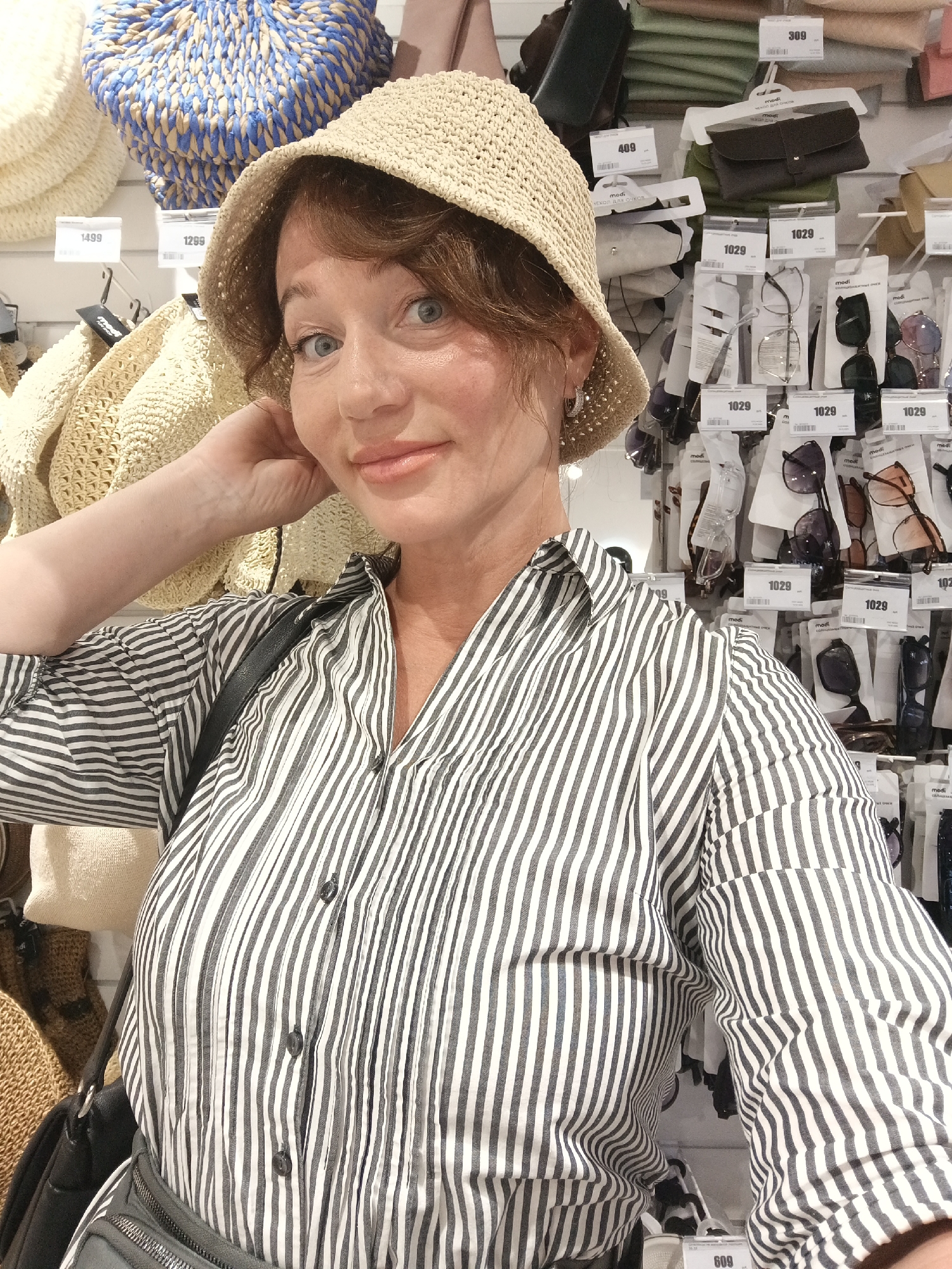 A woman with auburn hair, wearing a straw hat and a black and white striped shirt, poses for a selfie in a store. Behind her, shelves are stocked with various items, including hats and sunglasses. The image appears to have been taken in Sochi, Russia, possibly at the "Molodezhnyy" shopping center, given the presence of Russian price tags and the general retail environment. The woman is looking directly at the camera with a slight smile, her left arm resting on her head. The store appears to be well-lit and organized, with a focus on accessories and headwear.