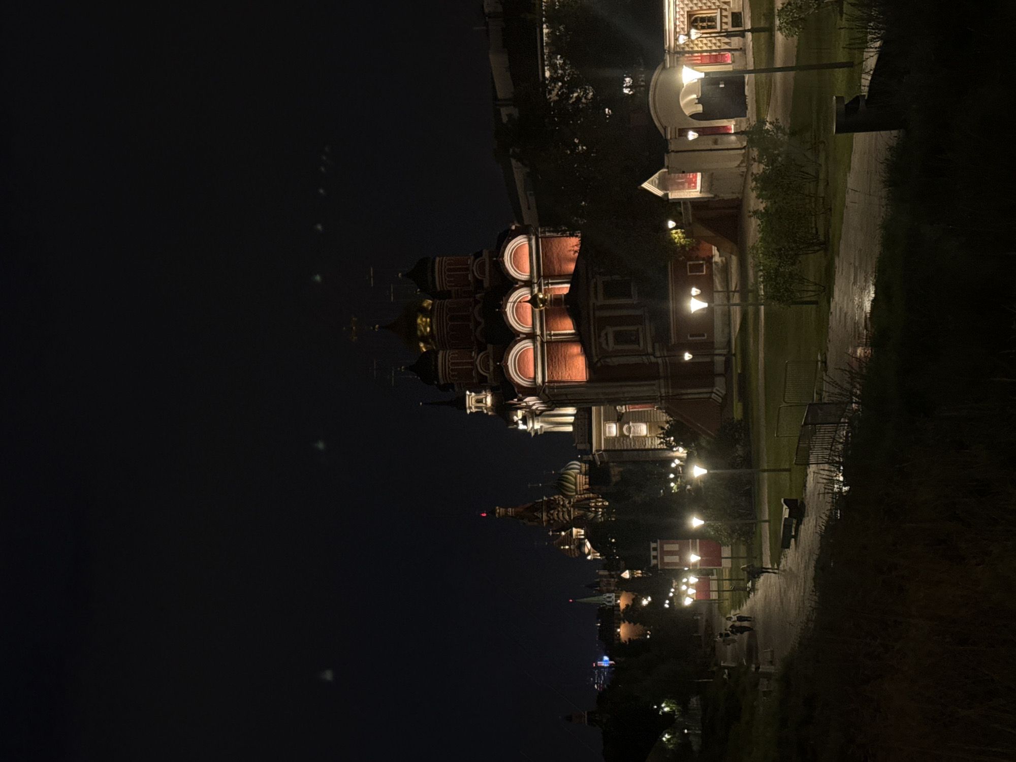 The image depicts a night scene in Moscow, Russia, likely Zaryadye Park, offering a view of historical architecture illuminated against the dark sky. In the background, a prominent red-brick building with golden domes and arched windows is brightly lit, possibly a church or part of the Kremlin complex. Several other spired structures with similar architectural styles are visible in the distance, also illuminated.
In the foreground, a paved pathway curves through a park area. The path is wet, suggesting recent rain, and is lined with lampposts that cast pools of light. Several people are walking along the path. Two individuals are seen walking together in the mid-ground, and a single person is walking further ahead. Benches are placed along the path, and there are grassy areas and trees surrounding the pathway. The overall atmosphere is serene and atmospheric, highlighting the illuminated historical landmarks of Moscow at night.