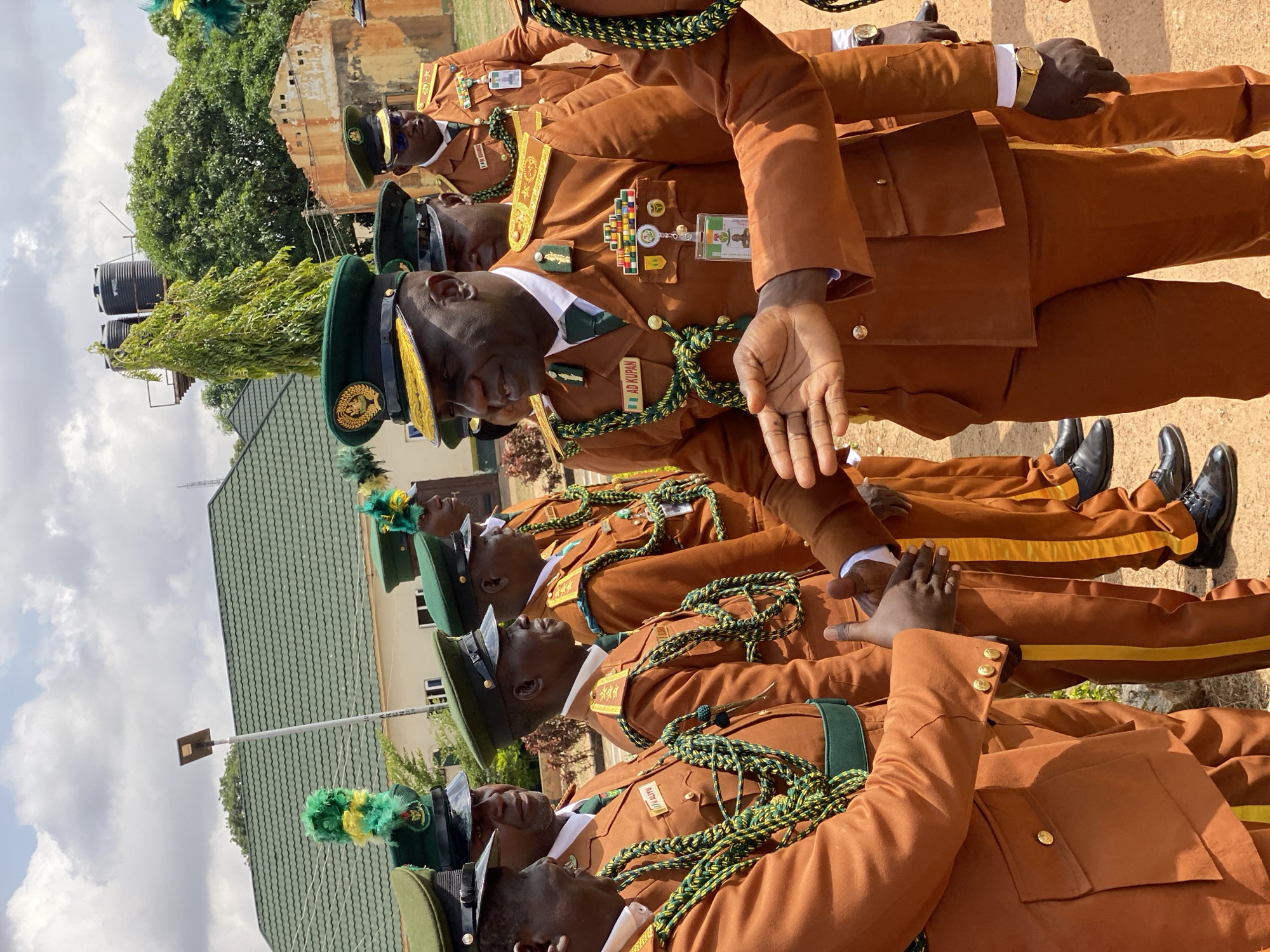 The image depicts a group of men in uniform standing in formation. The uniforms consist of brown jackets with gold buttons, white shirts, and green ties. They are also wearing green caps with gold emblems and green and yellow braided cords around their shoulders. Some of the men have medals or insignia on their jackets. One individual in the foreground is facing the camera and has his hand outstretched. The men appear to be participating in an event or ceremony. The background shows a building with a green roof, some trees, and a cloudy sky. The time of day appears to be daytime. There is some visible text on the uniforms, including "AD KUPAN" and "CATTIE III". The setting suggests an outdoor ceremonial event, possibly a parade or graduation, in Kaduna, Nigeria. The overall mood seems formal and disciplined.