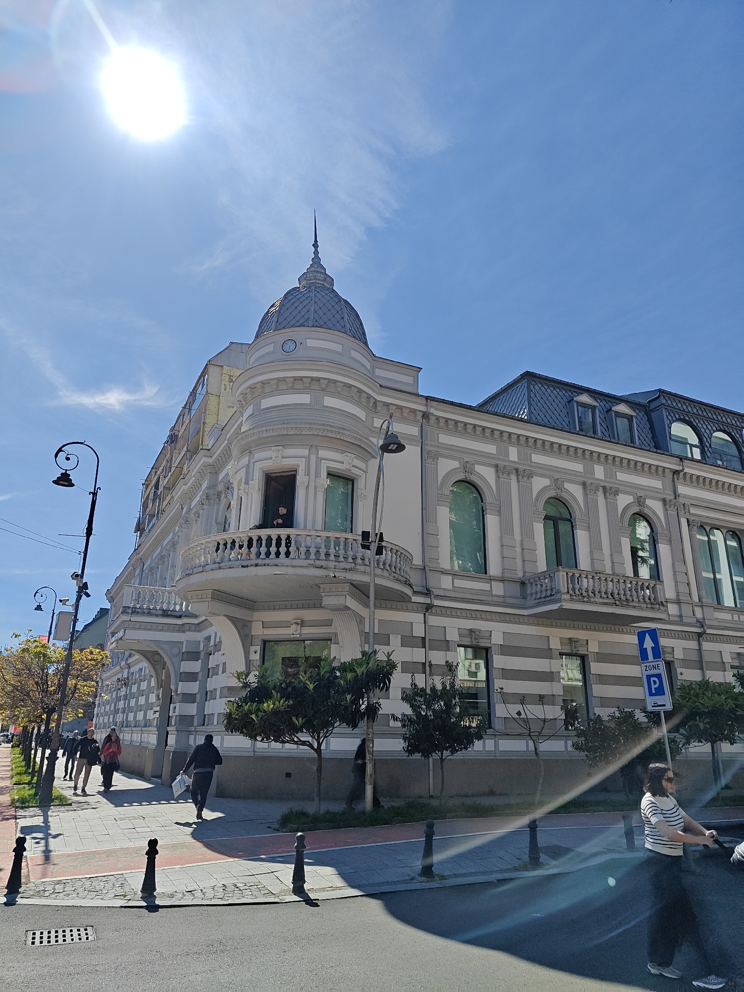 The photograph captures a street scene in Batumi, Georgia, under a bright, clear sky with the sun shining. A stately building, adorned with architectural details such as balconies, arched windows, and a domed turret, dominates the view. A person is visible on the balcony of the building. Pedestrians stroll along the sidewalk in front of the building, and a woman pushes a stroller across the street. Street lamps line the sidewalk, and small trees dot the streetscape. A blue parking sign is posted near the building. The street is shadowed in the foreground.