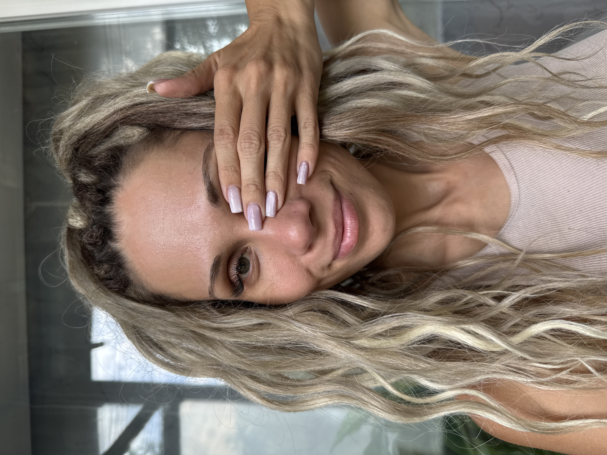 A medium close-up shot captures a woman with blonde, wavy hair and fair skin, who is covering her right eye with her left hand. She has on light-colored nail polish and is wearing a light-colored tank top. Her hair is pulled back in a high ponytail with some strands framing her face. She is smiling gently at the camera. The background appears to be an outdoor setting with some greenery and what looks like a window or glass door. The image was taken in Rusanovo, Russia.