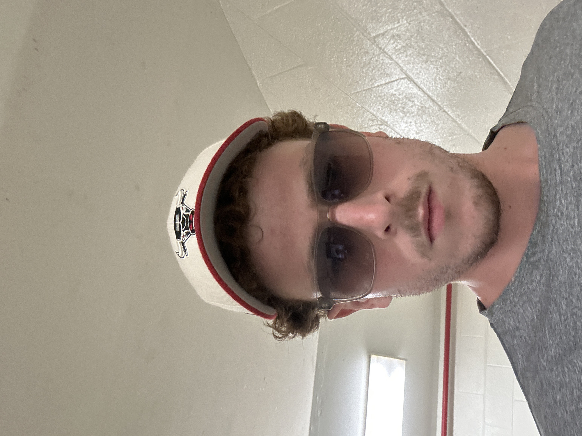 A medium-shot portrait of a man wearing sunglasses and a baseball cap with a logo, possibly taken in an indoor setting. The man is partially shown, with a gray t-shirt visible on the right side. The background features a white wall with rectangular tiles, suggesting an institutional or commercial space, with a door frame and possibly a thin red column also partially visible. Based on the prompt the location is inside of the city of Aventura, United States of America.