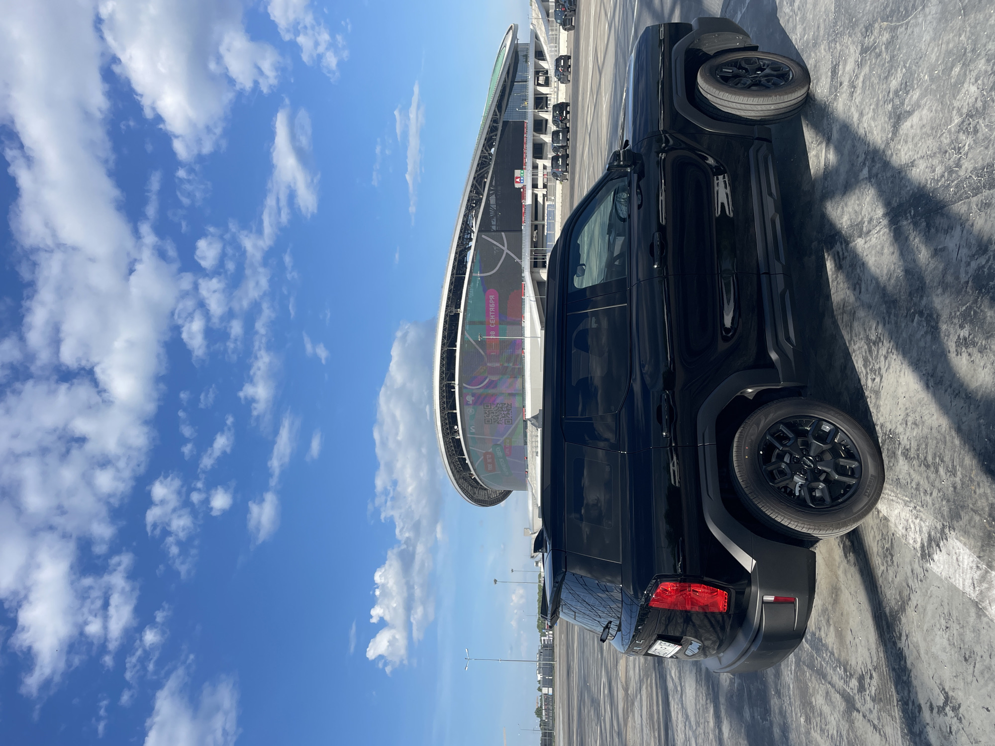 A black SUV is parked in an empty concrete lot in front of the Ak Bars Arena in Kazan, Russia. The stadium, with its distinctive curved roof and large LED screen displaying colorful graphics, is visible in the background. The sky is blue with scattered white clouds, and the sun casts shadows from the vehicle and streetlights onto the ground. Other cars are parked in the distance.