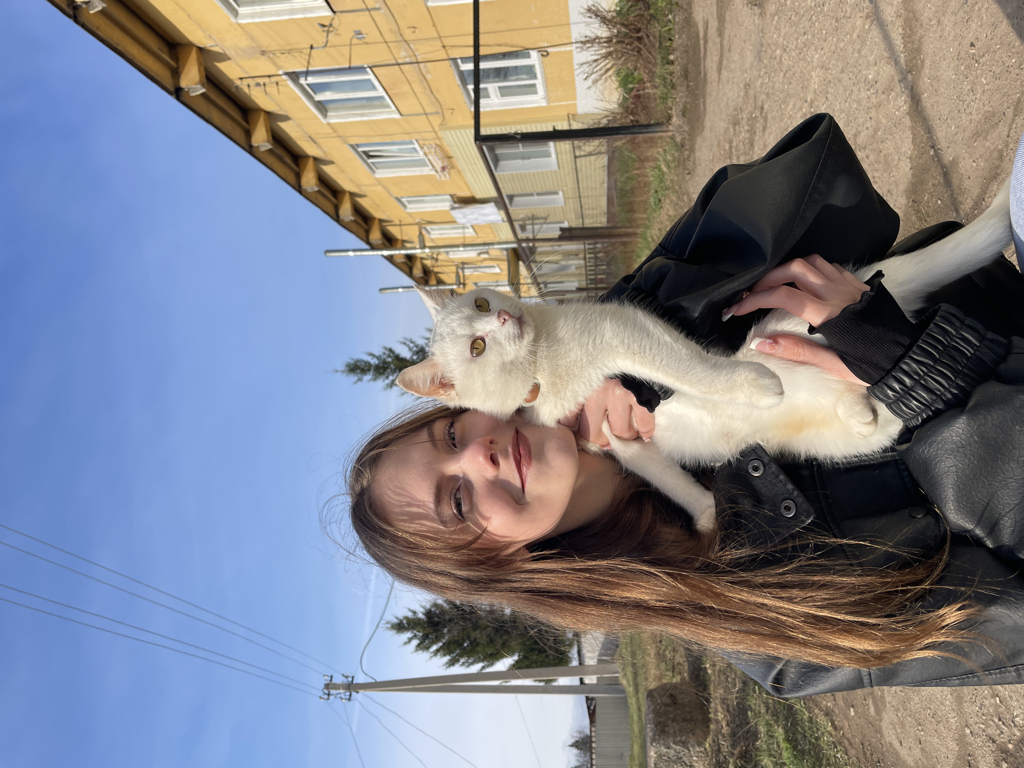 A young woman is pictured outdoors, holding a white cat against a backdrop of a residential building and a clear blue sky. The image appears to be a self-portrait or close-up photo, possibly taken on a sunny day.
The primary subjects are the woman and the cat. The woman has long, light brown hair, which falls over her shoulders. She is wearing a black jacket, possibly leather or faux leather. Her face is partially in shadow, but she has a subtle smile, indicating a calm or content mood. Her eyes are visible and look towards the viewer. The cat is predominantly white with hints of cream or light brown fur, and distinctive yellow-amber eyes that are wide open and looking directly at the camera. The woman holds the cat securely with both hands.
In the background, a multi-story building, painted in a faded yellow or light ochre color, is visible. It has several windows, some with white frames, suggesting it is a residential structure, possibly an apartment block. Architectural details, such as small triangular supports under the roofline, are also discernible. A clear, bright blue sky fills the upper portion of the frame, indicating sunny weather. A utility pole with several power lines stretches across the mid-ground. Sparse trees or bushes and an unpaved path or ground surface are also visible in the distance.
The activity is a posed photograph of the woman and her cat. The time of day appears to be daytime, likely late morning or early afternoon, given the bright, clear conditions and the quality of light. No visible text is present in the image. The setting suggests an outdoor residential area, consistent with the provided location context of Фоки, Russia.