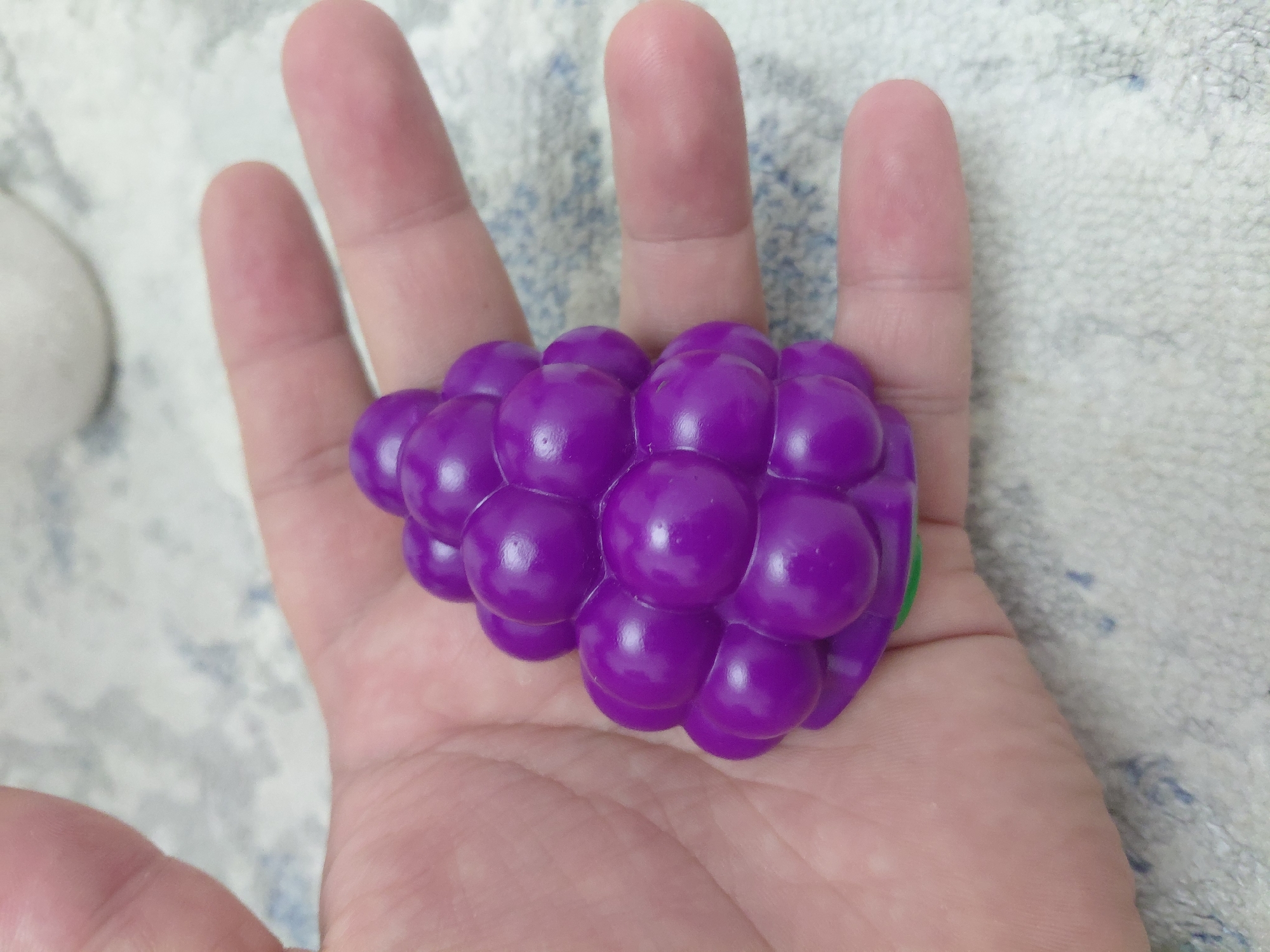 A close-up shot shows a person's hand holding a purple grape-shaped stress toy. The toy is composed of many small, round spheres clustered together, resembling a bunch of grapes. The hand is open, with the toy resting on the palm and fingers. The background is a blurry, light-colored textured surface, possibly a rug or towel. The image does not provide any information about the city of Aktau, Kazakhstan, or its specific location within the city.