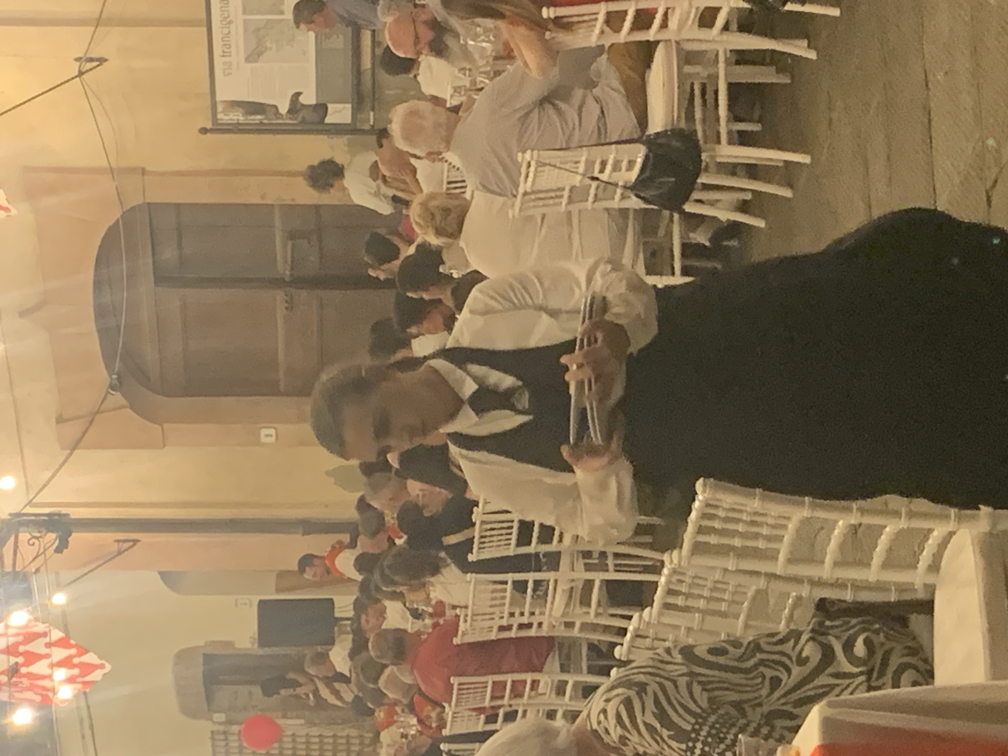The image depicts an outdoor evening gathering in San Quirico d'Orcia, Italy, likely at a restaurant or event space. In the foreground, a server wearing a white shirt and a dark apron holds a stack of plates. The background shows tables set with white chairs, where numerous people are seated, engaged in dining and conversation. The architecture suggests a historic Italian setting, with arched doorways and warm-toned walls. String lights are visible overhead, contributing to the ambient lighting. A red balloon is also noticeable in the distance. The scene conveys a lively and festive atmosphere.