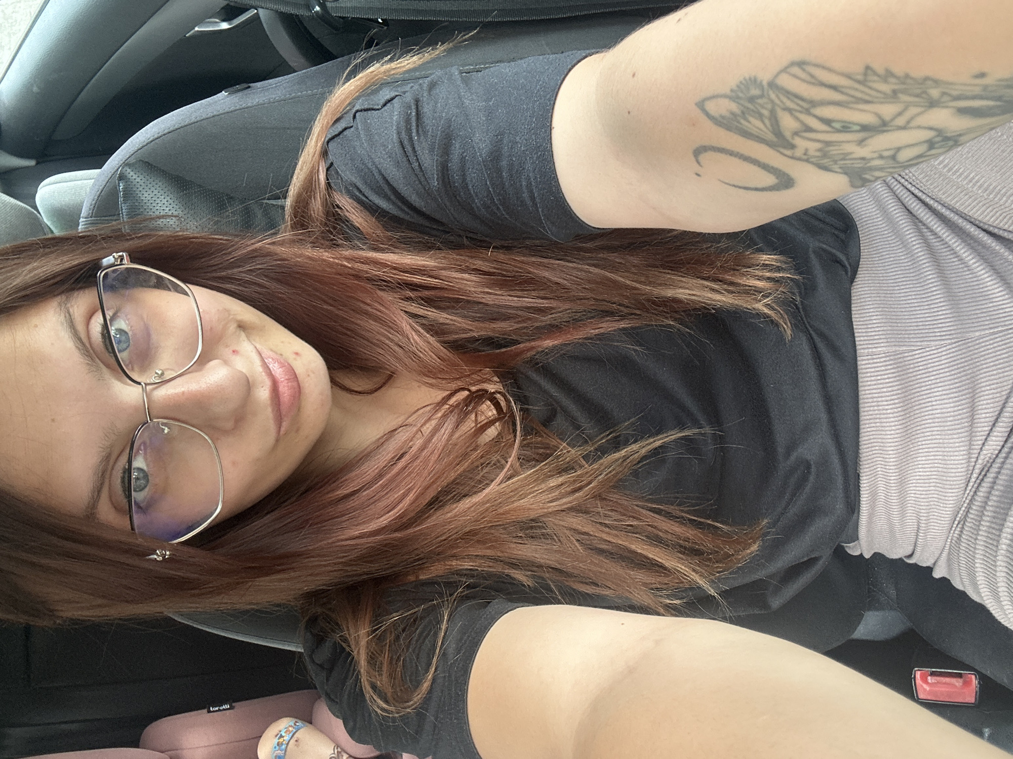 A young woman with long, brown, reddish-tinted hair and glasses is seated in a car. She is wearing a black t-shirt and light-colored, ribbed pants. She has a tattoo on her left arm. The car seat is visible behind her. There is a pink child car seat to her left. The overall scene appears to be indoors or in a shaded outdoor area, likely within the city of Tryavna, Bulgaria, based on the provided context.