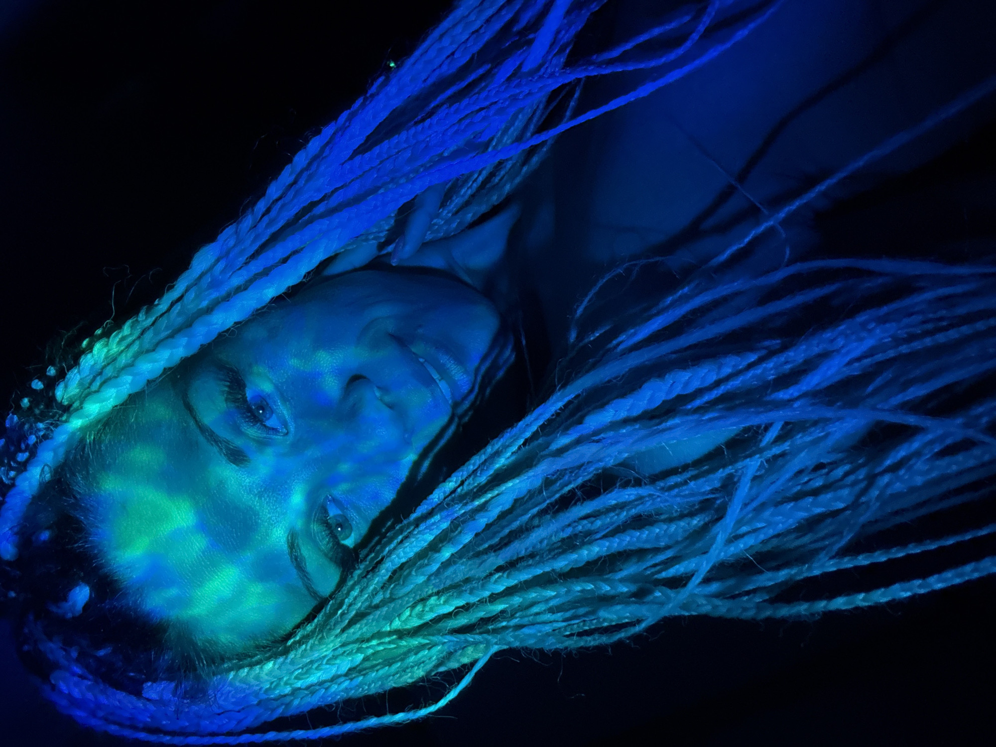 The image is a close-up, low-angle shot of a person's face and braided hair, illuminated by blue and green lights against a dark background. The person, with light-colored braids that extend out of the frame, has their face tilted and is looking towards the camera with a slight smile. The lighting creates a dappled effect on their forehead and cheeks, highlighting their eyes and eyebrows, and casting shadows. There are no discernible details about the location in the city of Rusanovo, Russia. The subject's clothing is not clearly visible.