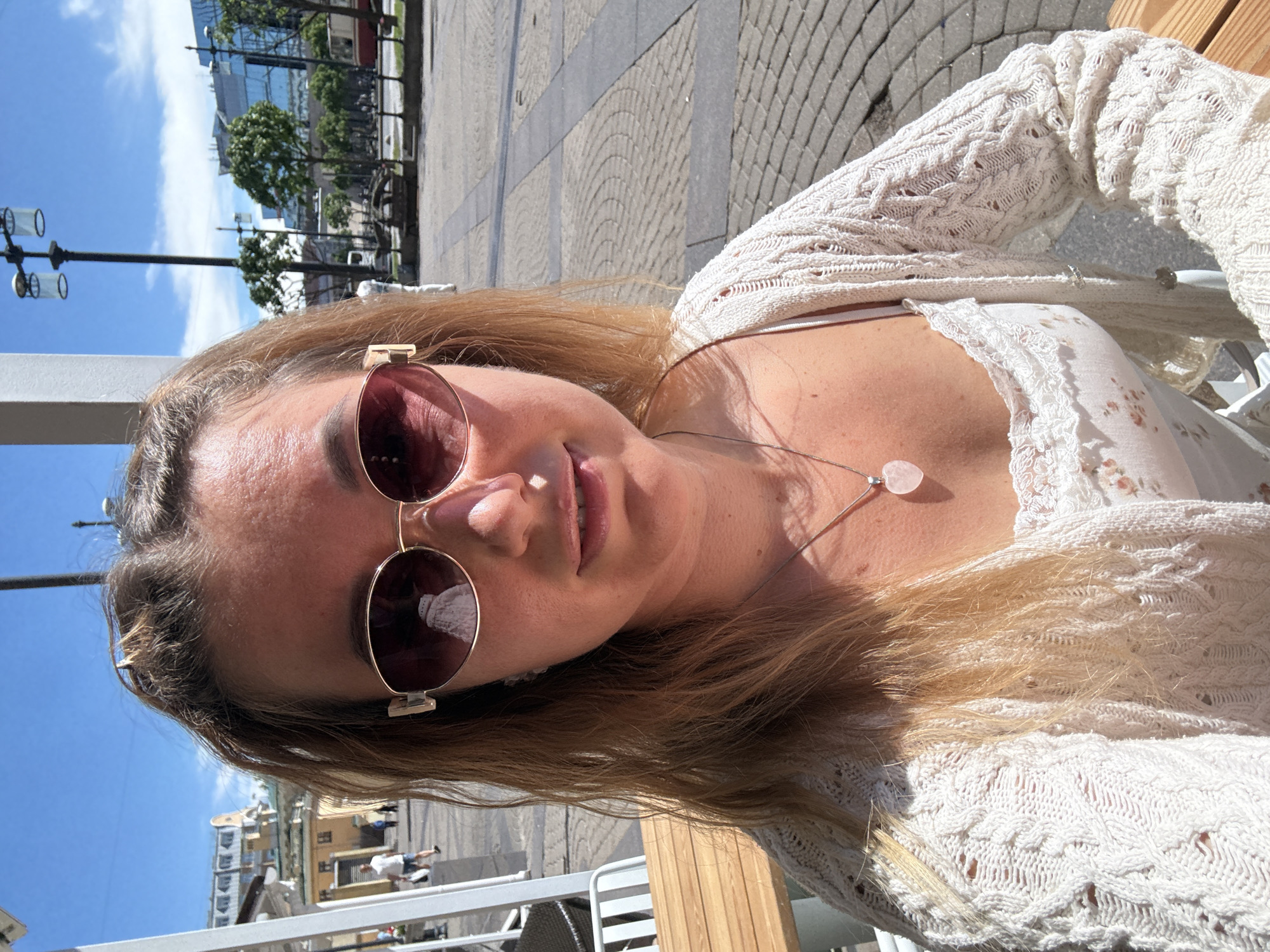 A woman with long, light brown hair, pulled back at the crown, is centrally positioned in a close-up outdoor photograph taken on a sunny day. She wears rose-tinted oval sunglasses with gold frames; a white architectural structure, possibly a dome or spire, is reflected in her left lens. Her expression is a slight smile. She is dressed in a cream-colored, open-knit crochet cardigan over a white top with a delicate pattern or lace trim, and a silver chain necklace with a light pink heart-shaped pendant rests on her chest. She appears to be seated at a light-toned wooden table, with parts of white metal chairs visible to her left.
The background depicts an urban outdoor setting. To the left, a white vertical pillar is visible, along with a yellow building featuring a green roof and multiple windows in the middle distance. Pedestrians, including a man in a white t-shirt and shorts, are walking in the background. To the right, a black lamppost with two cylindrical lights stands against a clear blue sky with minimal clouds. Further right, a modern building with a reflective glass facade is visible behind several trees. The ground is paved with interlocking stones, displaying both linear and circular patterns. A red and white vehicle is faintly discernible in the far distance on the right.