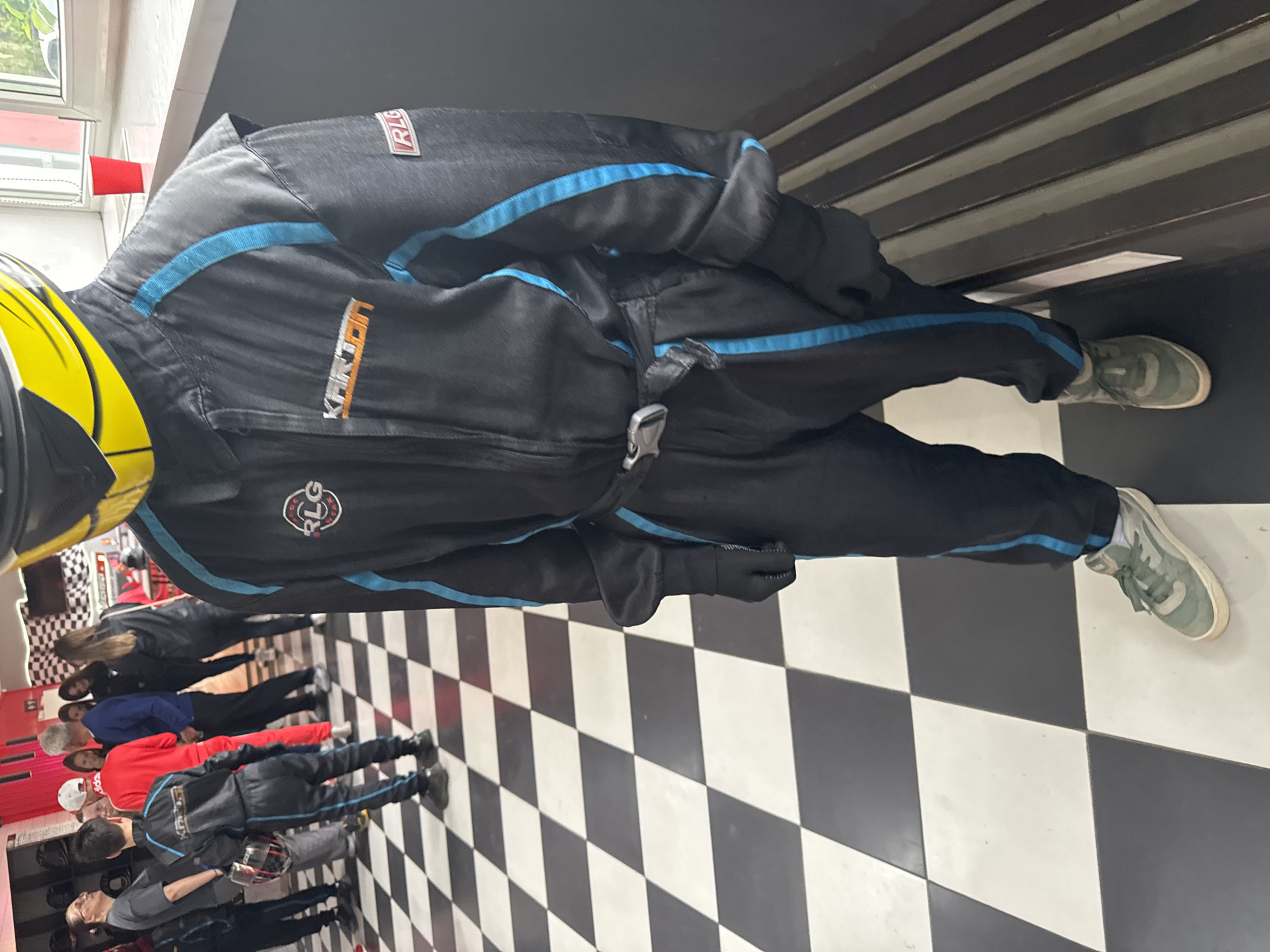 The image shows a person wearing a black racing suit with blue accents and a yellow helmet, standing on a black and white checkered floor. Several other people are standing in the background, and some are wearing similar racing suits. A red cup is visible in the upper left corner. The racing suit has "KARTODROM" printed on it and a logo that says "RLG." The location is Kartodrom Perm, in the city of Perm, Russia.