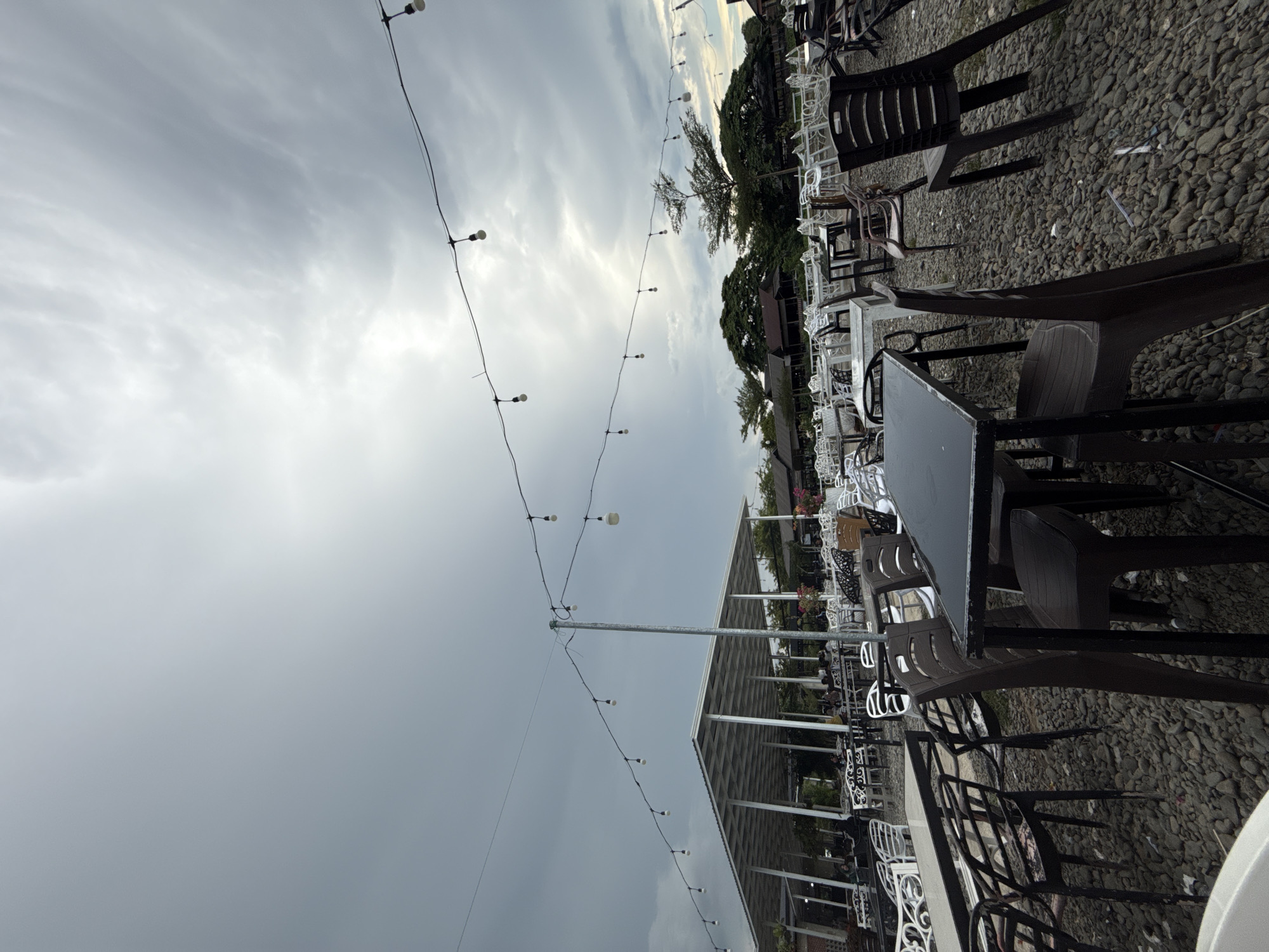 The image depicts an outdoor seating area of a restaurant or cafe, possibly in Cotmalem, Indonesia, under a cloudy sky. The ground is covered in small, gray stones. Numerous tables and chairs are arranged throughout the space, with a covered seating area visible in the background. String lights with exposed bulbs are hung across the scene. The tables are mostly dark, with some appearing to be metal-framed. The chairs are varied, with a mix of plastic and metal designs in dark colors. Some chairs are empty, while others appear occupied by people who are not clearly visible. The overall impression is that of a casual, open-air dining environment.