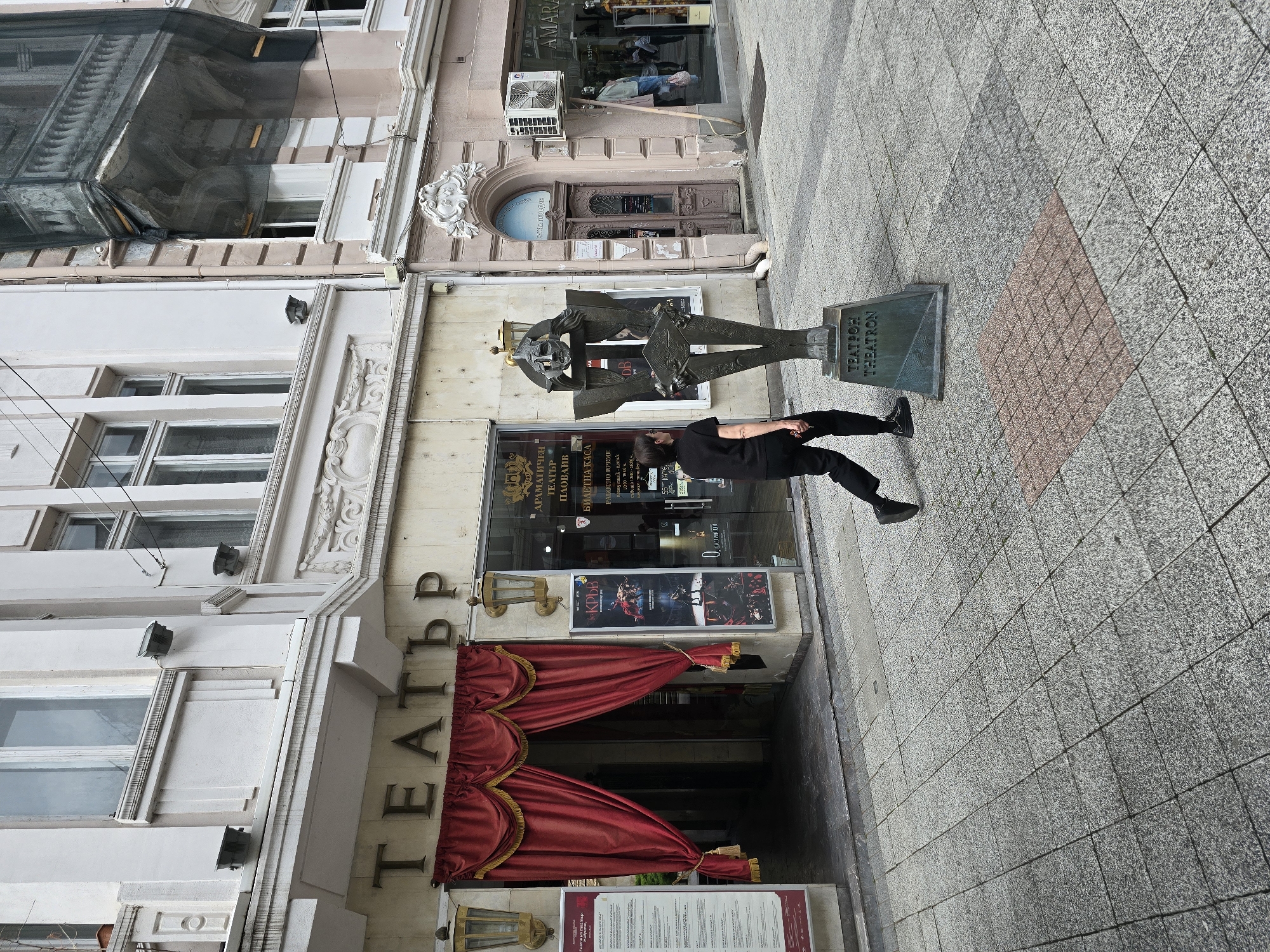 A street scene in an urban area, likely a city center, shows a person in black clothing interacting with a statue of a person in front of a building labeled "ТЕАТЪР," translated as "Theater," suggesting this is the entrance to a theater. The theater's entrance is marked by red curtains draped over the doorway and posters for the performance "КРЪВ," translated as "Blood." The theater is named ДРАМАТИЧЕН ТЕАТЪР ПЛОВДИВ, "Dramatichn Teatar Plovdiv", indicating it is located in Plovdiv. A ticket office window shows opening hours. The statue's base has the inscription "ТЕАТРОН THEATRON."