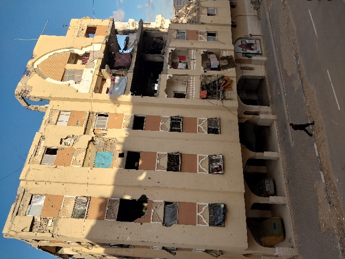 This image depicts a severely damaged multi-story building located in Khan Yunis, Palestinian Territory. The structure, predominantly beige in color, features at least six visible floors above a ground level characterized by arched open-air spaces. The building exhibits extensive damage, with numerous impact holes covering its facade and large sections of the upper right portion completely collapsed, exposing internal rooms and structural rebar. A prominent, partially destroyed decorative arched alcove is visible on the upper right. Multiple windows are broken, missing panes, or covered with various materials, including a blue tarp on the third floor from the bottom left. Balconies on the right side are structurally compromised, and laundry hangs from lines.
At ground level, the arched spaces serve as covered parking, with a green-covered vehicle and a silver sedan visible. A commercial storefront with a colorful graphic is partially visible on the far right. A paved sidewalk runs in front of the building, bordered by an asphalt road with a visible white dashed line. A single individual, dressed in dark clothing, walks from left to right on the sidewalk, casting a shadow. The sky above is clear blue, indicating daytime illumination, with shadows suggesting either morning or late afternoon. Debris is scattered along the base of the building and on the sidewalk.