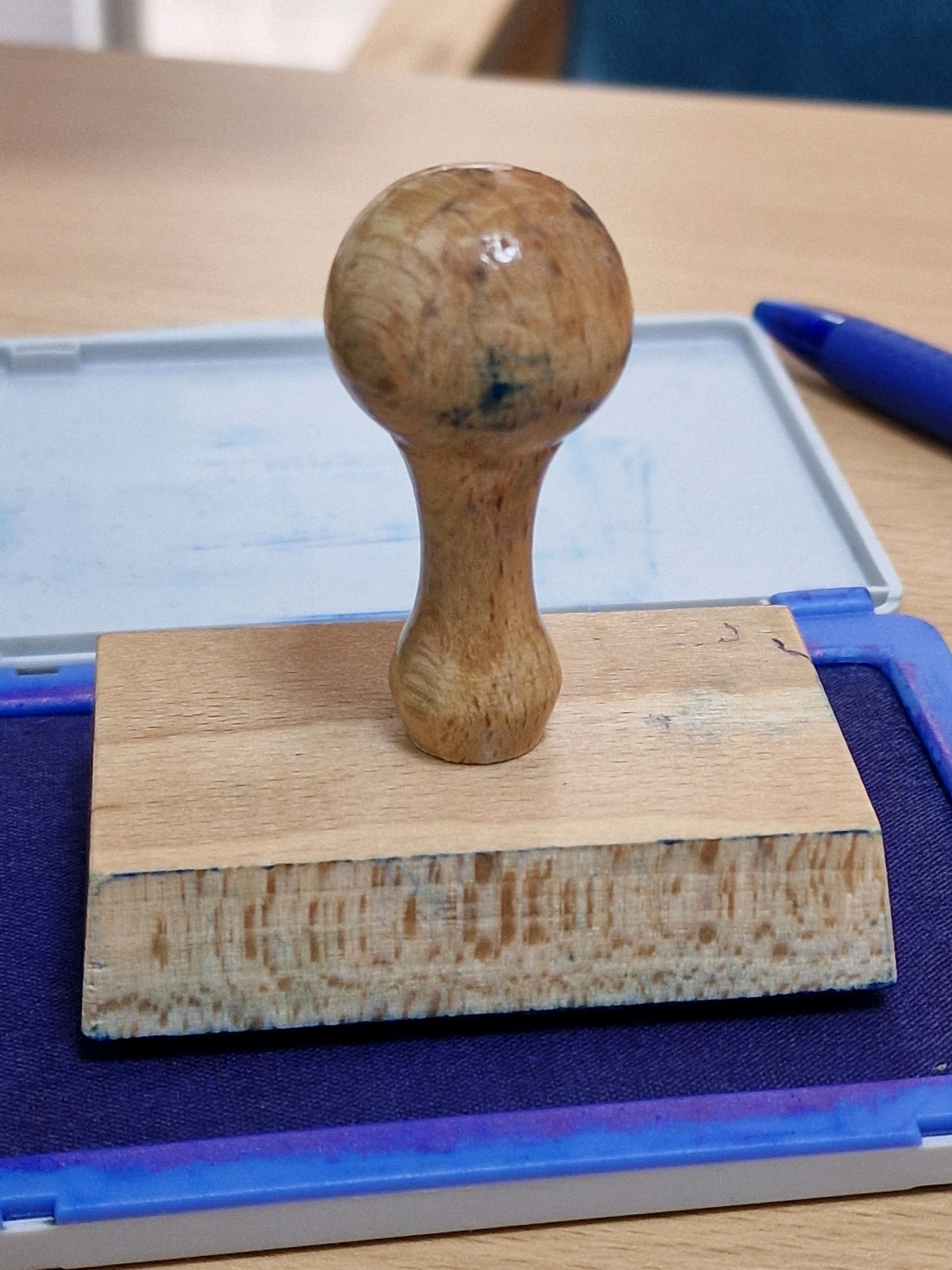 The image shows a close-up, high-angle shot of a wooden-handled rubber stamp resting on its wooden base, which is inside a blue ink pad. The ink pad is within a white and blue plastic casing. The stamp's wooden handle is rounded at the top and tapers down to the wooden base. The wood grain is visible on both the handle and the base. The ink pad itself is a dark blue color, and there are hints of purple where the ink has smudged or dried. A blue ballpoint pen is partially visible in the upper right corner of the frame, angled away from the stamp. The objects are placed on a light-brown wooden surface, likely a desk or table. The background is slightly out of focus, showing a lighter-colored surface behind the ink pad and a dark blue surface in the far background, possibly furniture. The lighting appears to be from above and slightly to the left, creating soft shadows. The image quality is sharp, with clear details of the textures and colors of the objects. This image was taken in the city of Travnik, Bosnia and Herzegovina.