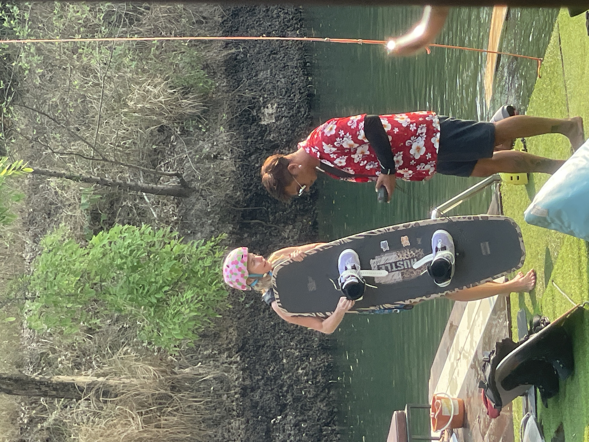 A woman wearing a pink polka dot helmet is being assisted onto a wakeboard on a dock. She is wearing wakeboarding bindings. A man wearing a red floral shirt is assisting her. The location is likely at or near the water in Tambon Nam Phrae, Thailand. The background includes trees, water, and a green artificial surface. A tow rope extends over the water.