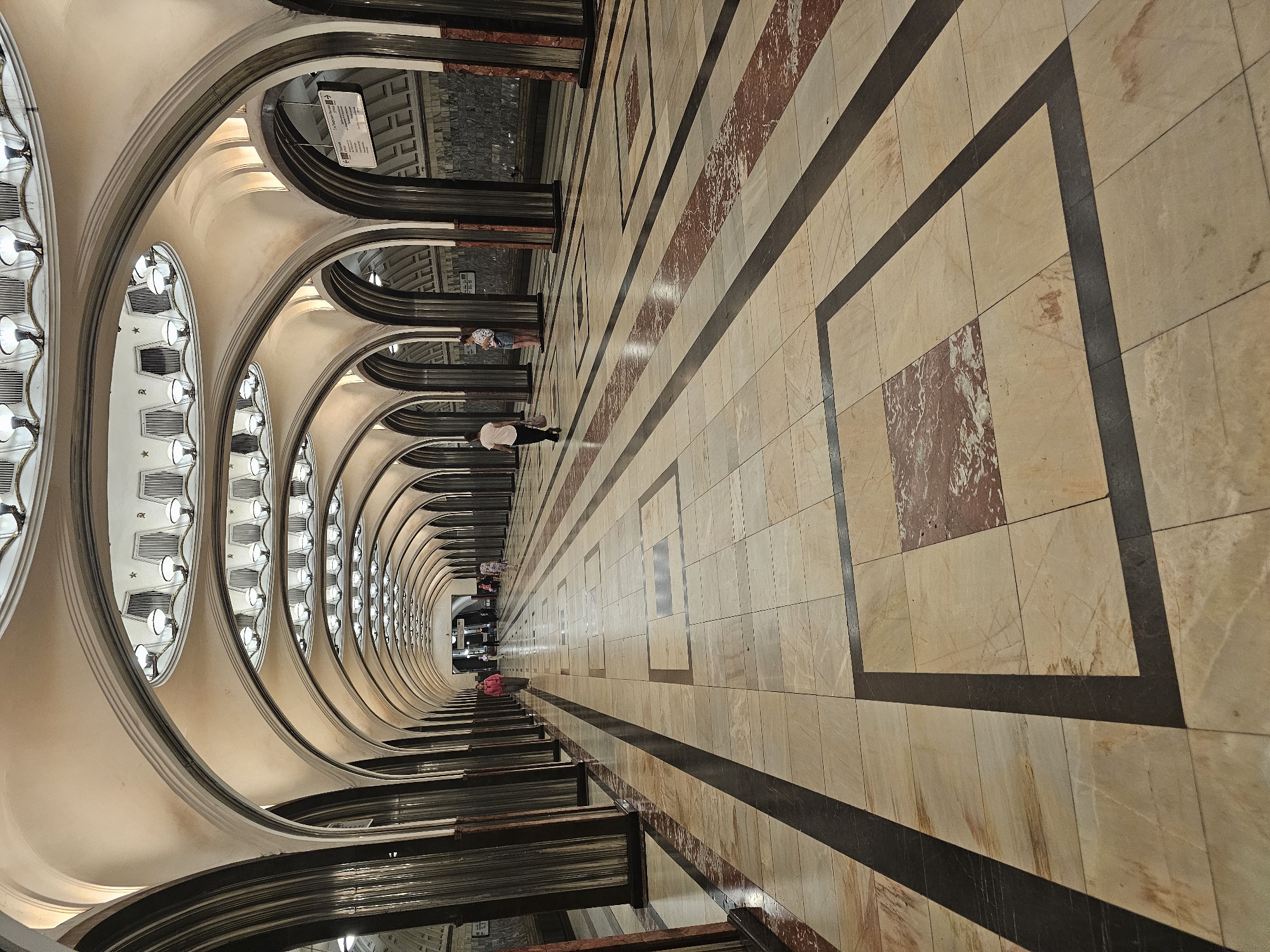 The image shows a wide, ornate hallway of a Moscow Metro station. The hallway features a series of arched alcoves with decorative lighting fixtures in the ceiling. The floor is covered in large, light-colored tiles with dark and reddish-brown accents, arranged in a geometric pattern. Dark, fluted columns line the sides of the hallway, supporting the arches.
There are several people visible, walking in both directions down the hallway. In the foreground, the floor is empty, drawing the eye towards the perspective of the station. In the middle ground, a person in a pink jacket is walking away from the camera, and a person in a white shirt and shorts is standing near a column on the right. Further down the hallway, other individuals are walking and standing, some near advertisements or signage. The overall impression is one of grandeur and spaciousness, characteristic of many Moscow Metro stations, which are known for their architectural significance.