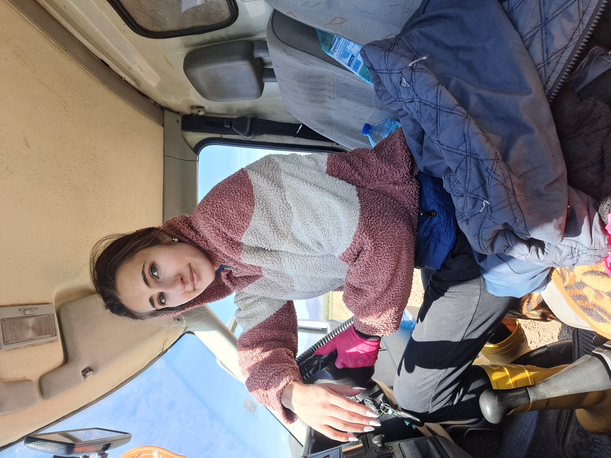 The image captures a young woman seated in the driver's seat of a vehicle, in Sofia, Bulgaria. She appears to be in her late teens or early twenties, with dark hair, green eyes, and a pleasant, calm expression as she looks directly at the camera. She is wearing a cozy-looking hooded jacket with distinct blocks of pink/mauve and cream/white fleece material. Her outfit includes black pants, and she has a bright pink glove on her right hand, while her left hand, adorned with white-painted nails, rests on the steering wheel. Keys are visible in the ignition.
The interior of the vehicle suggests it might be a van or a utilitarian truck, characterized by its beige-colored, somewhat aged cabin ceiling and simple dashboard controls. A sun visor and an overhead light fixture are visible. Through the front windshield, a bright blue sky with sparse clouds indicates clear, sunny daytime weather. On the passenger seat, a pile of clothes, including blue jackets and shirts, is visible, along with two plastic water bottles. One water bottle clearly displays the text "Bovinets" on its label. The scene conveys a candid moment, possibly during a break or just before starting a journey.