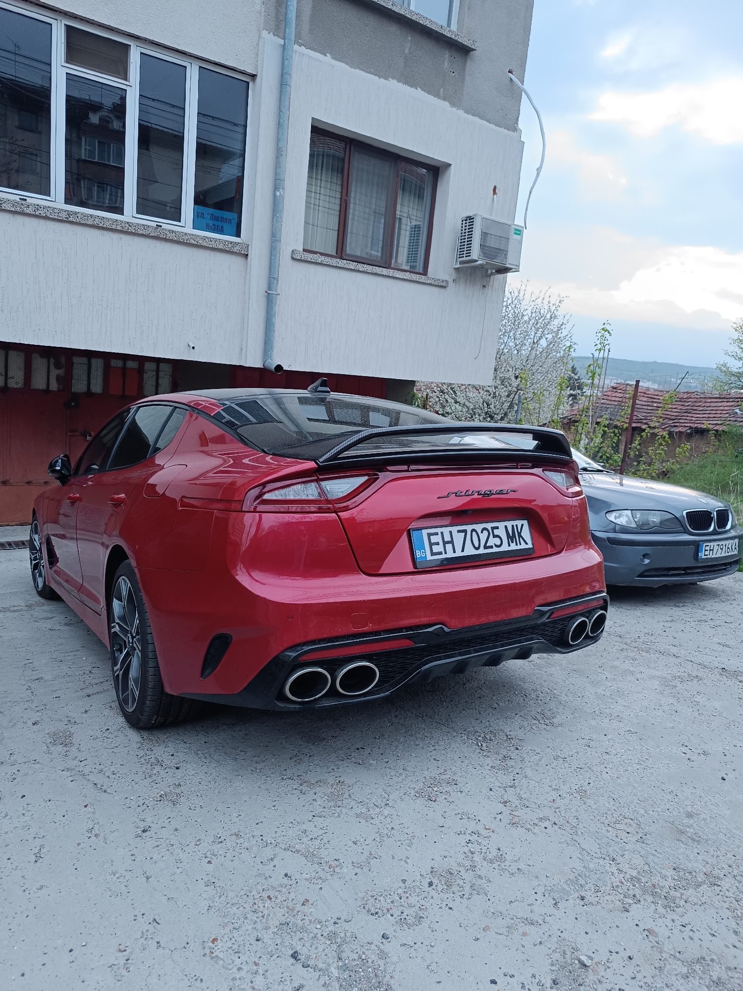 A detailed forensic description of the image is as follows:
The image captures a red Kia Stinger parked on a gravel surface in front of a building. The rear of the car is prominently displayed, showcasing the "Stinger" logo, Bulgarian license plate "EH 7025 MK", and quad exhaust pipes. To the right of the Stinger is a gray BMW with the license plate "EH 7916 KA".
The building behind the cars is a two-story structure with a light-colored facade. Visible features include windows with white frames, a metal drainpipe, and an air conditioning unit mounted on the wall. A sign on one of the windows reads "ул. "Люляк" №36A", indicating the location is on "Lilac" Street, number 36A.
In the background, there's a glimpse of a more rural landscape, with other buildings and greenery visible in the distance. The sky is overcast, casting a muted light over the scene.