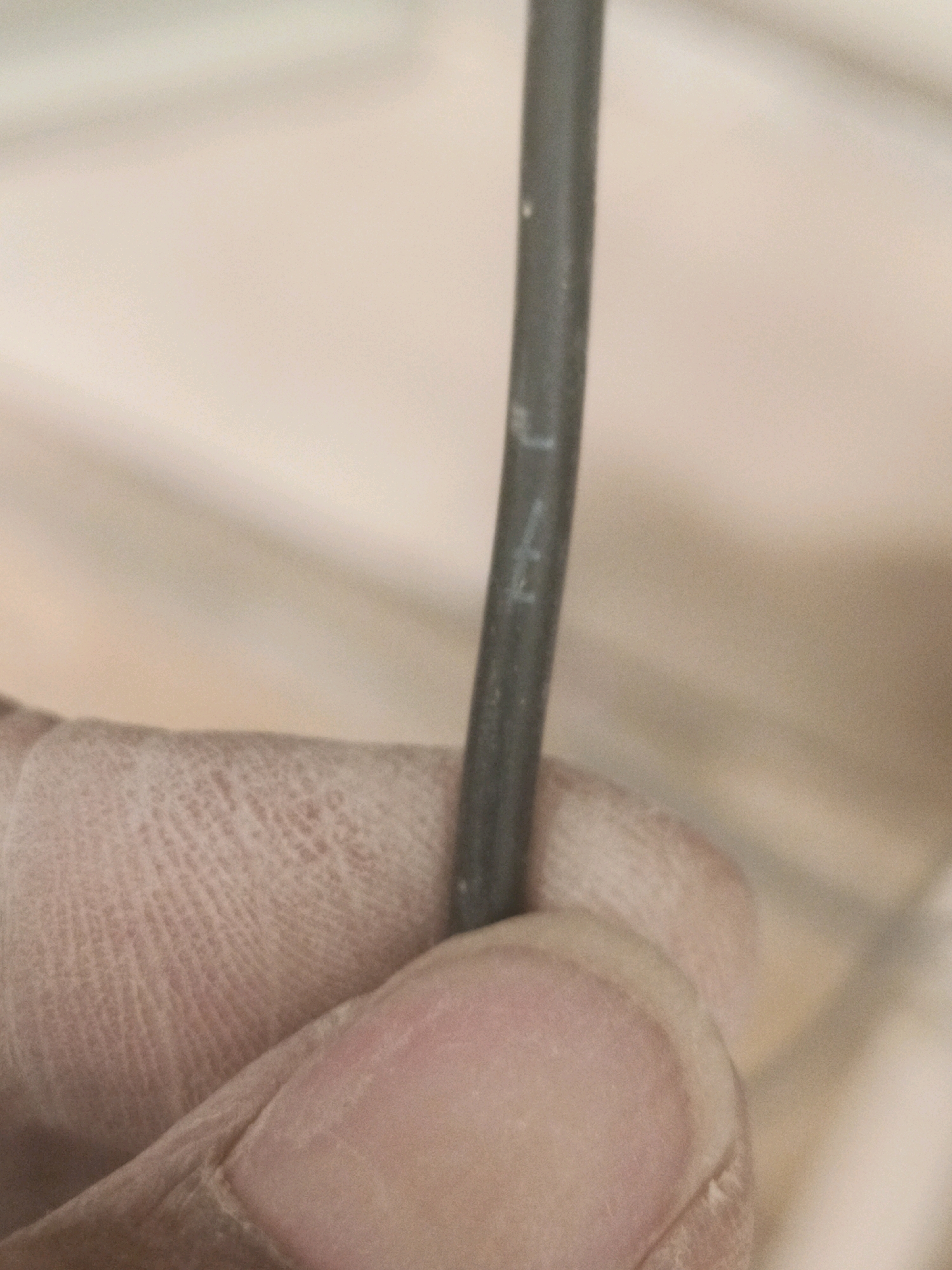 A close-up shot shows a grey cable being held by a person's hand, which is partially visible in the lower left corner. The cable has faint markings on it, possibly numbers or letters. The background is out of focus and appears to be a wall or surface with a light, neutral color. The image does not contain any discernible features that would allow for its localization to the city of Lebaniwaras, Indonesia, or any other specific geographic location.