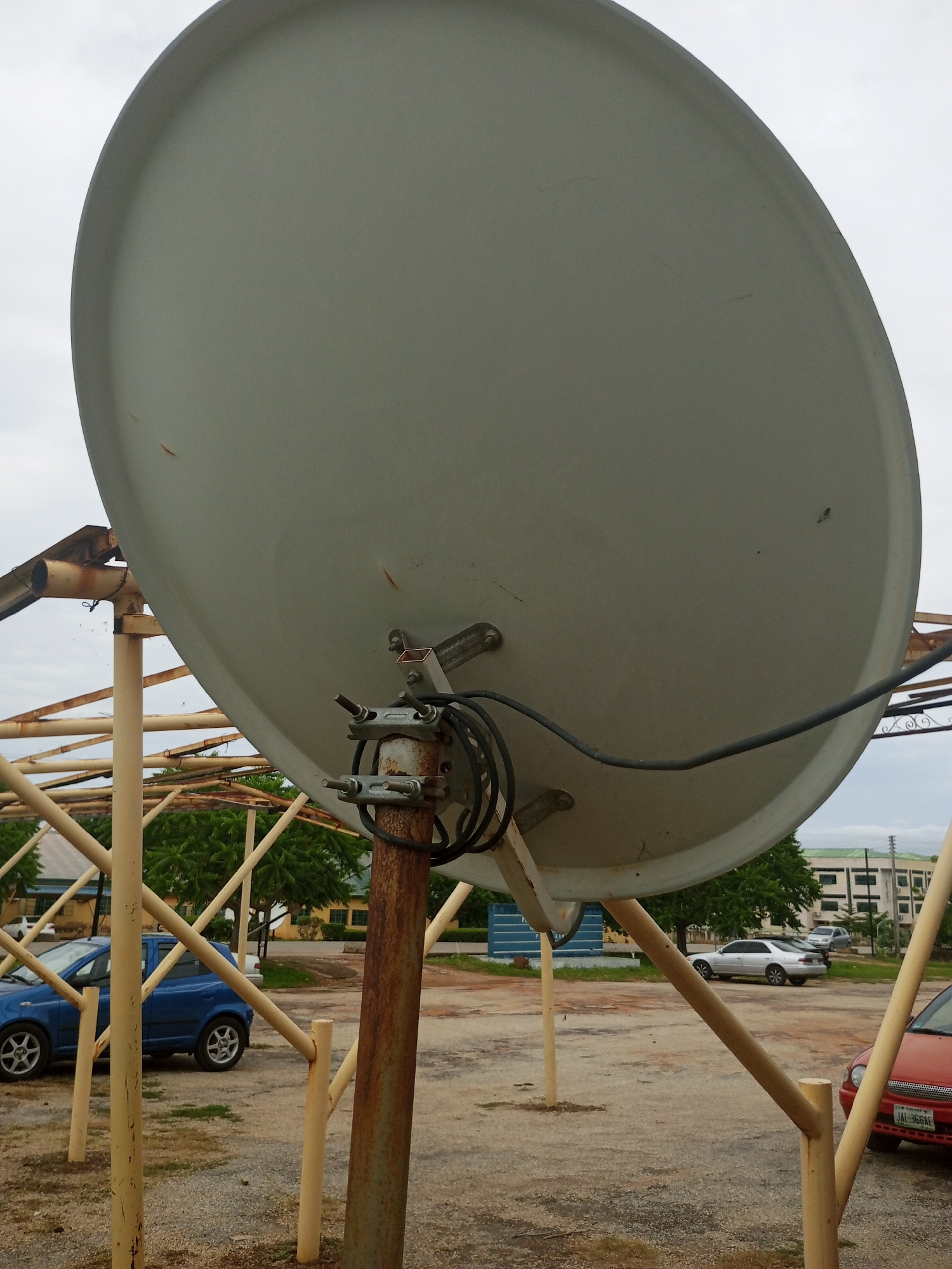 A large, light gray satellite dish dominates the foreground of this outdoor shot, angled upwards against a bright, overcast sky. The dish is mounted on a rusty metal pole and connected to a complex network of yellowish, weathered metal scaffolding that forms an open, gazebo-like structure. A black cable snakes from the dish, secured by metal clamps to the mounting pole.
In the mid-ground, a vibrant blue hatchback car is parked behind the scaffolding. Further back, several other cars are visible, including a silver sedan and a red sedan. In the background, there are low-rise buildings with green roofs and a line of trees with lush green foliage.
The scene appears to be set in an open area, possibly a parking lot or a yard, with gravel and dirt covering the ground. The cloudy sky suggests it could be a humid day in Jalingo, Nigeria, where the image was taken. There is no apparent activity or event taking place, with the focus being on the satellite dish and its support structure.
A license plate is partially visible on the red car in the lower right corner of the image, with the text "JAL 366AE". The overall impression is one of an ordinary day in a semi-urban or rural setting in Nigeria, with functional, perhaps aging, infrastructure.