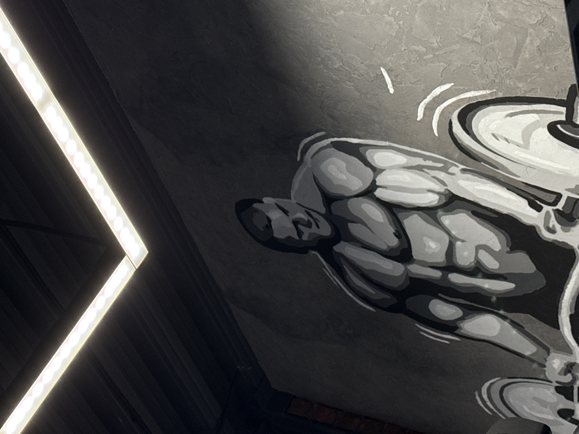 The image shows a wall mural in a gym, possibly located in Votkinsk, Russia. The mural depicts a male weightlifter in grayscale, performing a deadlift with a barbell. The background is a textured gray wall. A bright, linear LED light fixture is visible on the left side of the frame, positioned diagonally. The style of the mural is stylized and graphic.