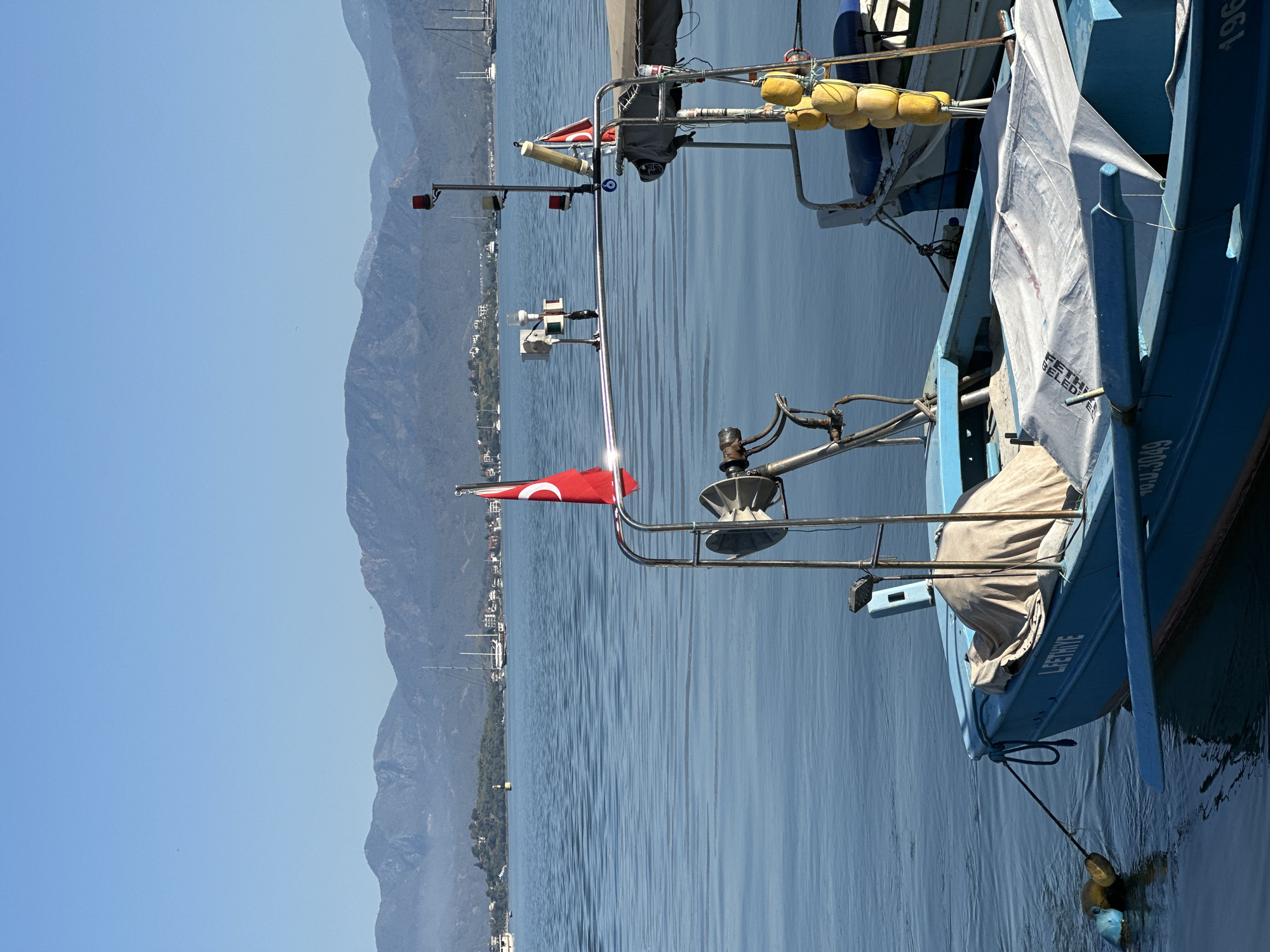 This image captures a serene maritime scene in Fethiye, Turkey, focusing on a blue boat set against a backdrop of calm waters and distant mountains.
The main subject is a light blue boat, likely a fishing vessel or small utility boat, seen from its stern and starboard side. A metal railing structure along the boat's back features various attachments, including yellow and blue buoys, red navigation lights, and a winch-like mechanism. A red Turkish flag, bearing a white crescent moon and star, proudly flies from this railing. Additionally, a blue evil eye charm (Nazar boncuğu) hangs from the railing, a cultural symbol common in Turkey. A white canvas tarp covers part of the boat, with the visible text "FETHİYE" and "SELED" printed on it. Further text on the blue hull includes "LEENHE" and the registration number "BM.3049," with a partial number "196" also visible.
The boat rests in tranquil, blue water that subtly mirrors the clear, bright blue sky. It appears to be stationary, tethered by a rope to a yellow buoy in the water. In the background, a range of mountains stretches along the coastline, with scattered white buildings nestled at their base. Masts of other boats are discernible in the distance, indicating a nearby harbor or bay.
The scene unfolds under a clear, sunny sky, suggesting bright daylight, likely morning or early afternoon. The weather is calm and clear, contributing to the peaceful atmosphere. No people are visible in the image. The overall composition conveys a sense of quiet coastal life in the identified location of Fethiye, Turkey.