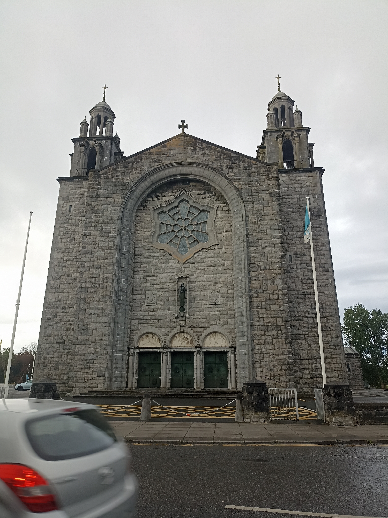 The image captures the facade of the Galway Cathedral, a prominent landmark in Galway City, Ireland. The cathedral is constructed of grey stone blocks and features two tall bell towers with crosses on top. A large rose window is centrally located above the entrance, which consists of three large green doors. A statue of a religious figure is positioned above the doors. In front of the cathedral, a sidewalk is visible with yellow cross-hatching markings and a low stone wall. A white metal gate is also present. A tall flagpole with a blue and white flag stands to the right of the entrance. The foreground shows a silver car partially blurred due to motion, driving on an asphalt road. The sky is overcast. To the right of the cathedral, some trees are visible.