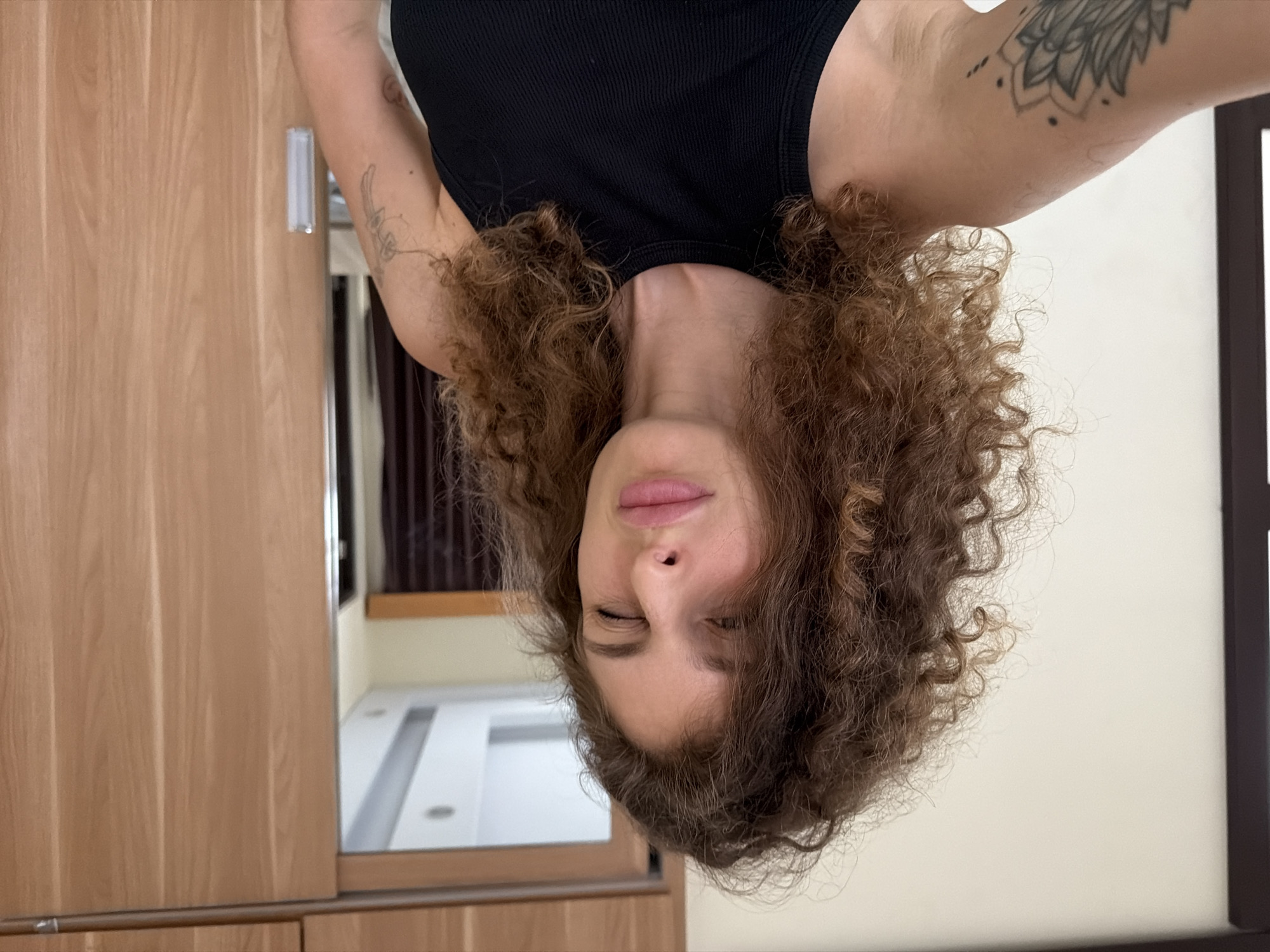 A woman with curly brown hair and a black tank top is winking at the camera. She has pink lips and her eyes are closed on one side. She is standing in front of a closet with wooden doors, which is likely in her home or a hotel room in North Kuta, Indonesia. There is a tattoo of a mandala on her left arm.