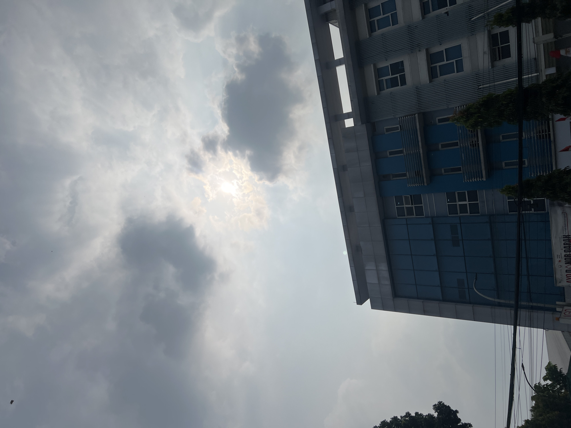 The image captures a cloudy sky with the sun peeking through the clouds, casting a bright glow. In the foreground, a modern building with a gray and blue facade stands tall. The building has multiple floors with windows and balconies. Some trees are visible on the left and right sides of the building. The sky is dominated by large, gray clouds, suggesting an overcast or partly cloudy day. The building appears to be located in an urban or semi-urban area, given the presence of utility lines in the lower portion of the image. Based on the provided context, the location is identified as Citeureup, Indonesia. There are no people visible in the frame, and no discernible actions are taking place. The overall scene is static and focused on the architecture and the sky.