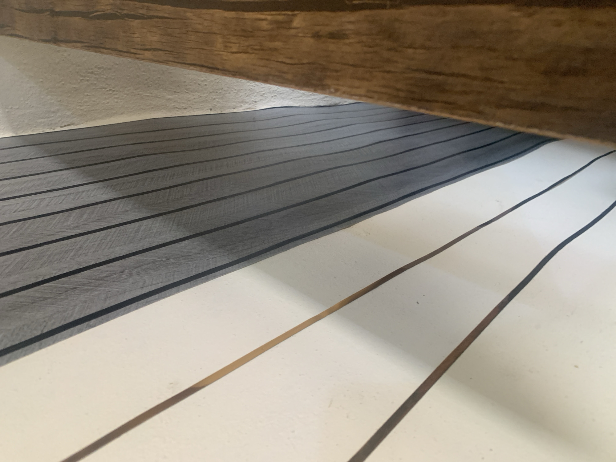 The image is a close-up, angled shot of a wall decorated with vertical elements. On the left, a white surface is adorned with thin, dark lines and wider, slightly darker brown lines that appear to be inset or painted on. These lines are spaced unevenly and run diagonally from the lower left to the upper right. Adjacent to the white surface is a section of grey wallpaper with a subtle wood grain pattern, also featuring thin, dark vertical lines. To the right of the grey wallpaper is a roughly textured, light-colored wall, and further to the right, a thick, rough-hewn wooden beam or post is visible, positioned at an angle and occupying the right side of the frame. The lighting suggests an indoor environment, possibly a staircase or hallway given the diagonal lines and angled perspective. The scene is static and devoid of any human presence or activity. The location is identified as Jauro Yinu, Nigeria.