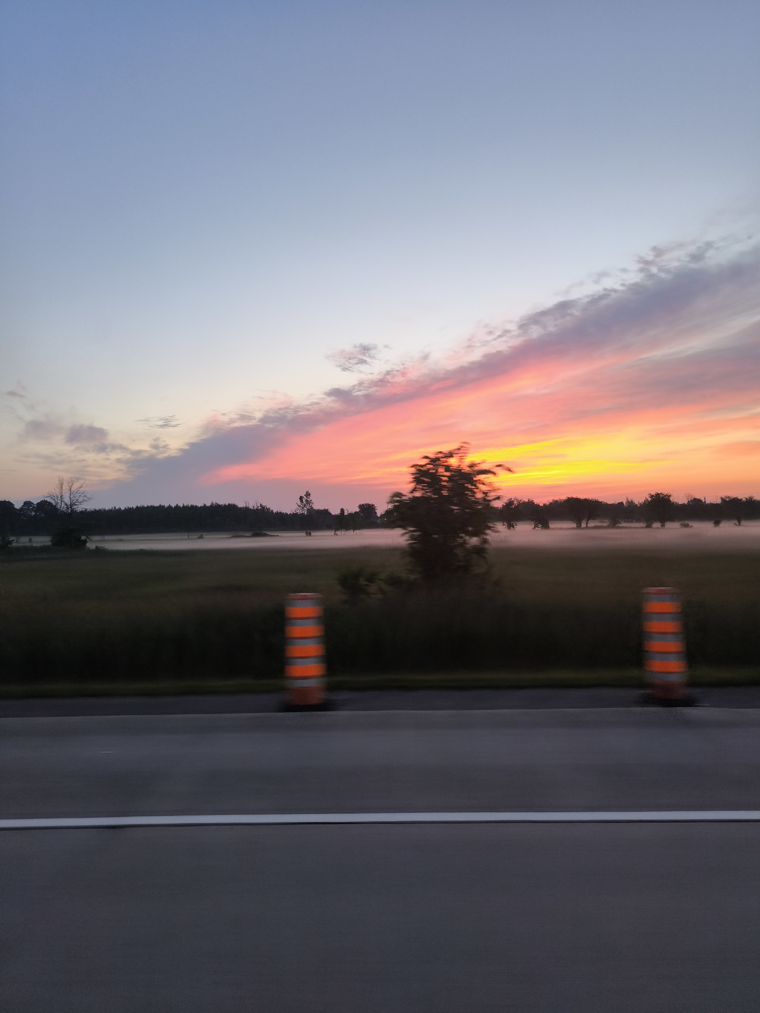 The image depicts a roadside view of a rural landscape at dusk. The sky is a gradient of soft blues transitioning to vibrant oranges and pinks, illuminated by the setting sun. Wispy clouds are painted with these sunset colors. In the midground, a field is covered in a low-lying mist that partially obscures a line of trees and silhouetted structures in the distance. The foreground features a section of asphalt road, distinguished by a white line. Two orange and white striped traffic cones are positioned on the shoulder of the road, indicating some form of traffic management or construction. The overall impression is one of tranquility and the natural beauty of twilight in a rural setting. The scene is likely captured from a moving vehicle, given the slight motion blur evident in the trees and cones. The location in Mirabel, Canada, suggests a setting characteristic of the Quebec countryside.