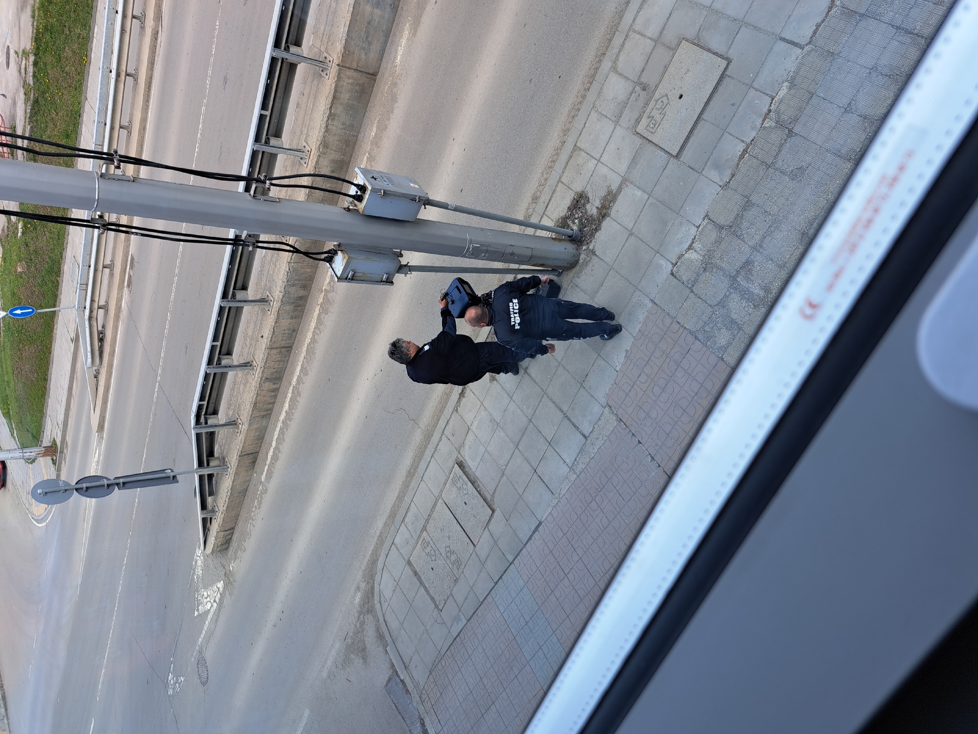 The image, taken from an elevated, oblique perspective, shows two men on a tiled sidewalk next to a street in Sofia, Bulgaria. The lighting suggests it is daytime, possibly late afternoon or an overcast day.
One man, wearing a dark uniform with "TRAFFIC POLICE" written in white on his back, is kneeling at the base of a grey street light pole. He appears focused on a blue device or bag placed on the ground beside him. Standing next to him is another man with grey hair, wearing a dark jacket, who is leaning towards the pole, observing or assisting.
The scene includes a wide asphalt road with white lane markings, bordered by a concrete barrier and a metallic safety guardrail. A tall, grey street light pole, equipped with several black electrical cables and two grey utility boxes, stands prominently on the sidewalk. The sidewalk itself is paved with a mix of grey and patterned tiles, featuring a square metal utility cover. A blue circular sign with a white left-pointing arrow is visible near a grassy area adjacent to the road, indicating a directional cue. A blurry, light-colored structural element, possibly part of a building or vehicle, occupies the right foreground, partially obscuring the sidewalk and displaying indistinct text that includes a 'CE' mark. The activity appears to be an inspection or work related to the street pole or electrical equipment by the traffic police and an accompanying individual.