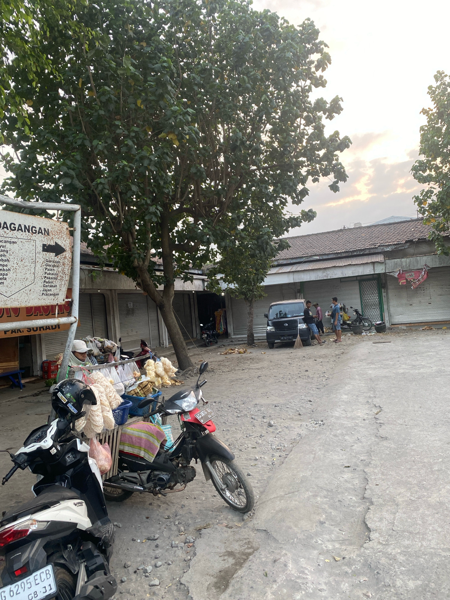 This image depicts an outdoor scene in Gurah, Indonesia, likely a market or a roadside area. In the foreground, two motorcycles are parked. The motorcycle on the left is white and black, and its license plate reads "G 6295 ECB 08-31". The motorcycle on the right is black and red, and its license plate is "AG 3674 GI 04-27". A person wearing a white cap and a blue shirt is standing behind the motorcycles, next to a stand laden with bags of snacks and what appear to be crackers. Behind the motorcycles and the vendor, a line of shuttered storefronts forms the background. A dark minivan is parked in front of one of these storefronts, and three men are standing near it. One man is holding a broom. The ground is unpaved and dusty. Trees with dense green foliage are visible, casting shadows on the scene. The sky above is overcast with some light breaking through, suggesting either early morning or late afternoon. A sign with text in Indonesian, including "DAGANGAN" (merchandise) and "PAK. SURADI" (Mr. Suradi), is partially visible on the left side of the image. The overall impression is one of a commercial area preparing for or winding down operations.