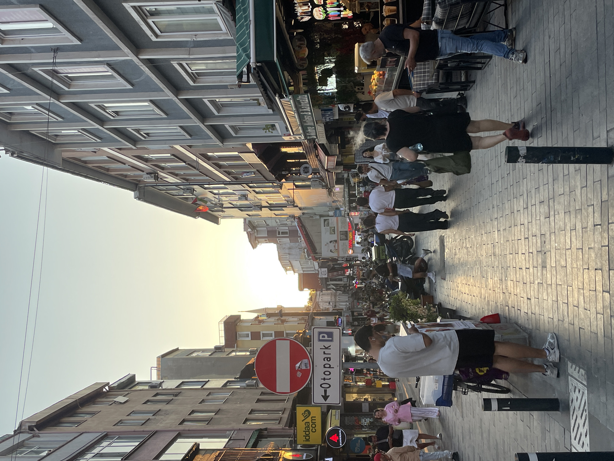 The image depicts a street scene in Kadıköy, Turkey, likely in the evening, as indicated by the warm, low-angled sunlight casting long shadows and illuminating the sky with soft, golden hues. The street is lined with multi-story buildings featuring apartment windows and shopfronts. Numerous people are walking along the pedestrianized street, some appearing to be locals and others tourists.
In the foreground, a man in a white t-shirt and black shorts stands looking at his phone. Further down the street, various individuals are seen. A woman in a black dress walks away from the camera, carrying a green bag. Another man, wearing a black t-shirt and blue jeans, stands with his phone in hand. A woman in a pink striped outfit and a pink headscarf walks towards the camera, and a man in a tan shirt and white shorts is also present.
The street is paved with cobblestones. On the left side of the street, there are signs for businesses, including "iddaa.com" and a circular red and white "no entry" sign with a white horizontal bar. A directional sign with an arrow and the word "Otopark" (Turkish for parking) is also visible, indicating a parking area to the left. Shopfronts display merchandise, and some have awnings providing shade. In the background, a minaret of a mosque is visible rising above the buildings. The overall atmosphere is one of a busy, commercial street in a city.